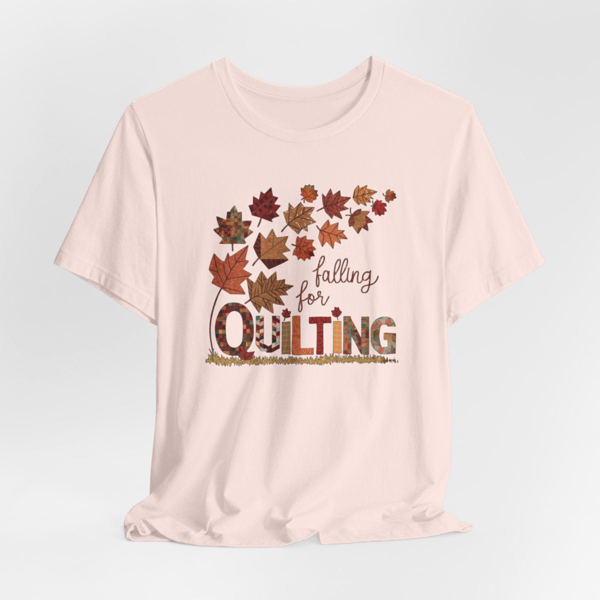 A Soft Pink thanksgiving quilting t-shirt featuring autumn leaves with various quilting patterns inside. Text reads "falling for QUILTING" with letters in patchwork style. Warm fall colors and quilting motifs combine in a seasonal design.