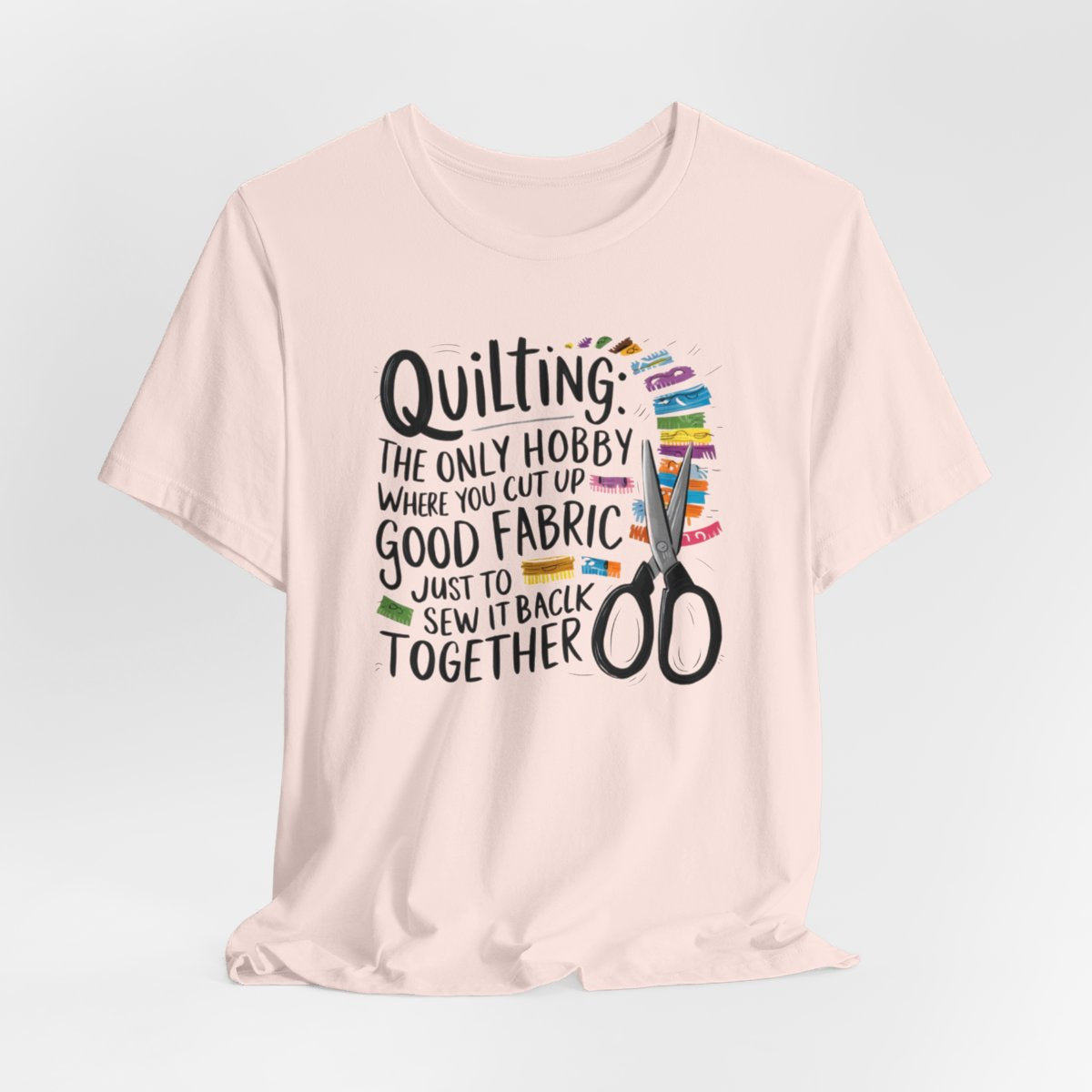 A Soft Pink Quilting-themed T-Shirt with text "Quilting: The Only Hobby Where You Cut Up Good Fabric Just to Sew it Back Together" surrounded by colorful fabric scraps and scissors.