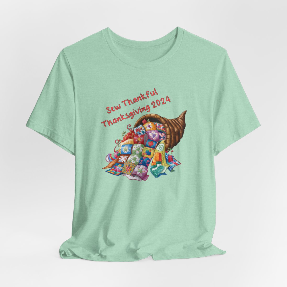 A Heather Mint thanksgiving quilting t-shirt featuring a colorful quilted cornucopia overflowing with various quilt patterns and designs. Text reads "Sew Thankful Thanksgiving 2024" in red. Vibrant illustration combines quilting motifs with Thanksgiving imagery.