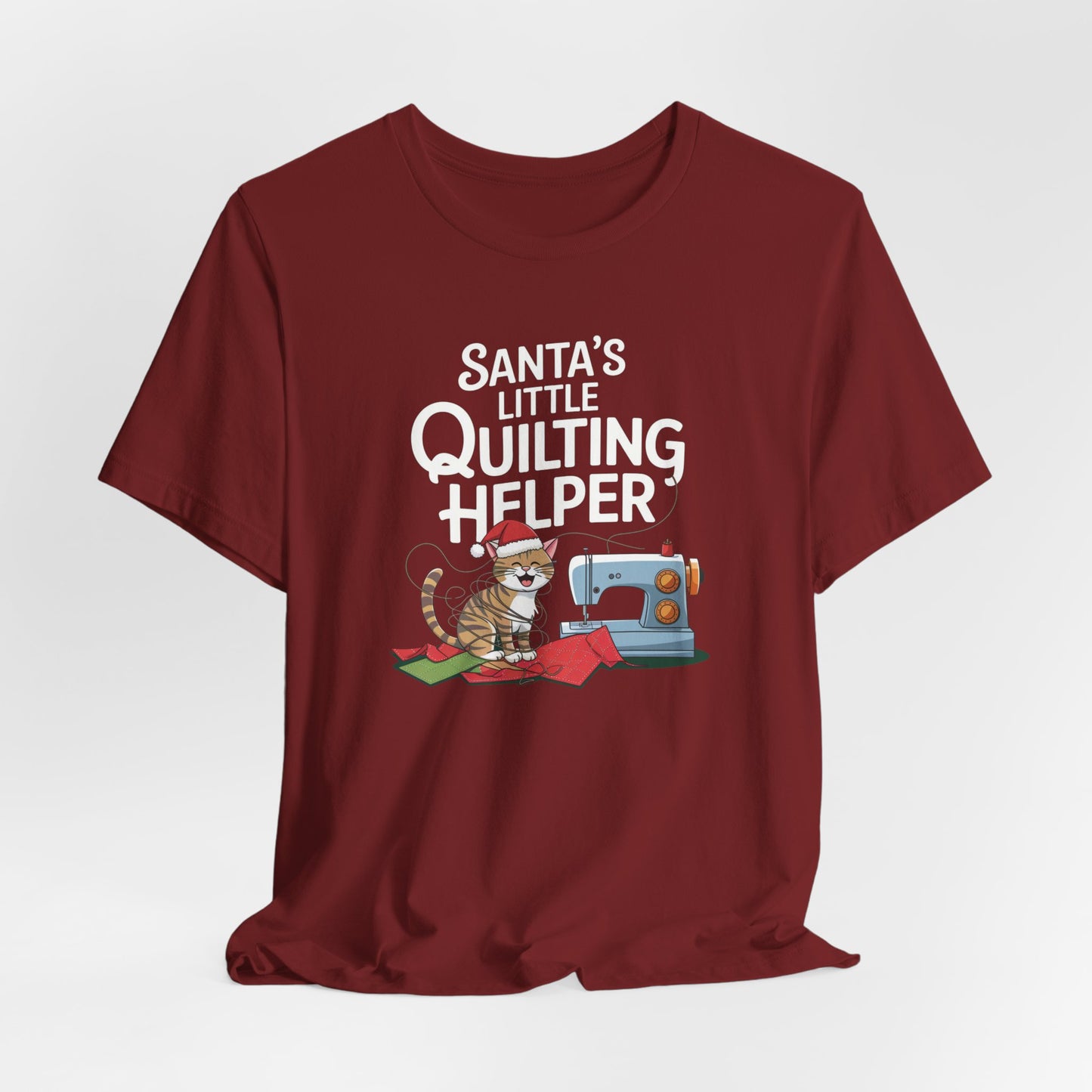 A Cardinal christmas quilting t-shirt featuring a playful cat wearing a Santa hat is tangled in thread next to a sewing machine, with the phrase "Santa's Little Quilting Helper" above, combining quilting and holiday humor.