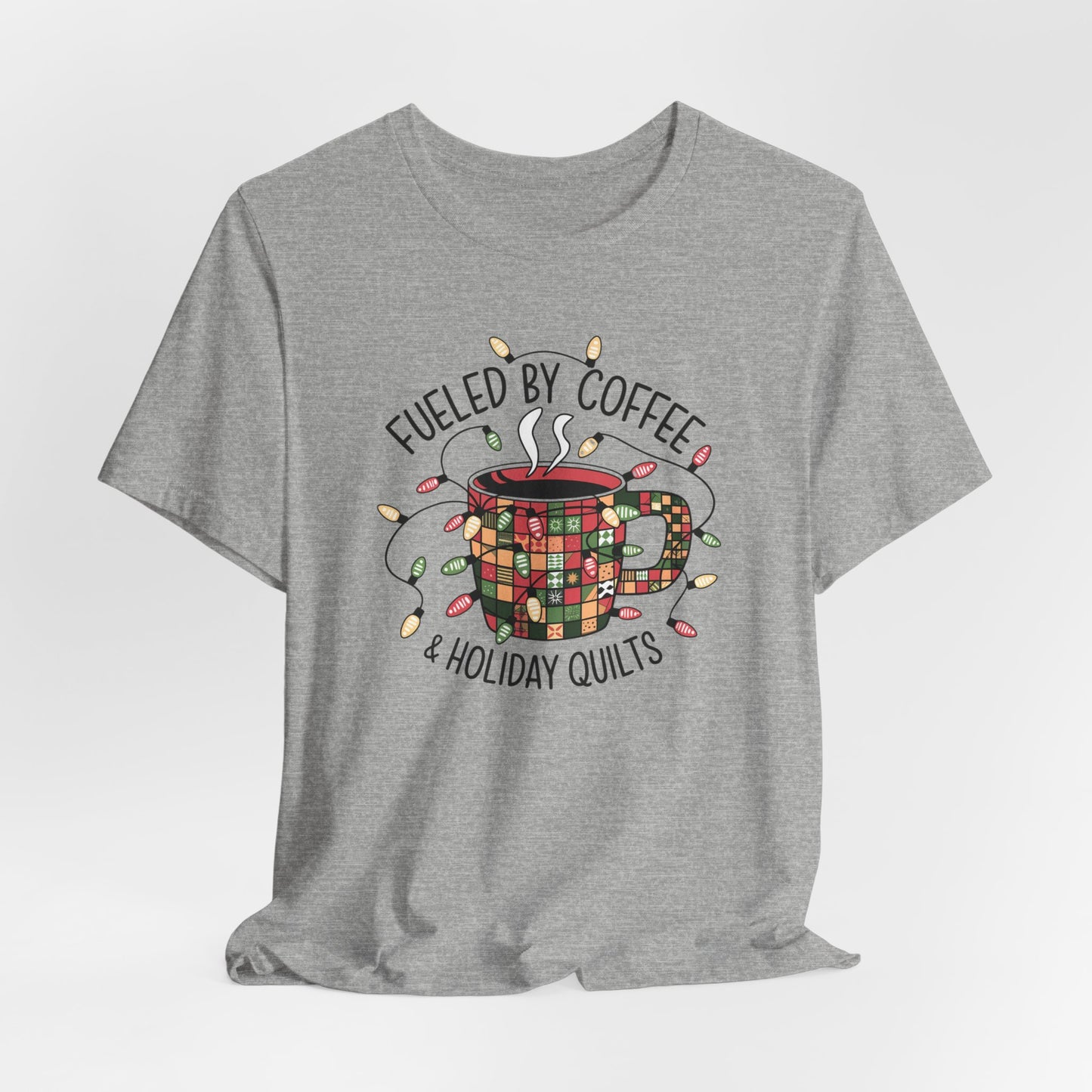 An Athletic Heather christmas quilting t-shirt featuring a cozy coffee mug covered in patchwork quilt patterns, wrapped in festive holiday lights, with the phrase "Fueled by Coffee & Holiday Quilts" surrounding the design.