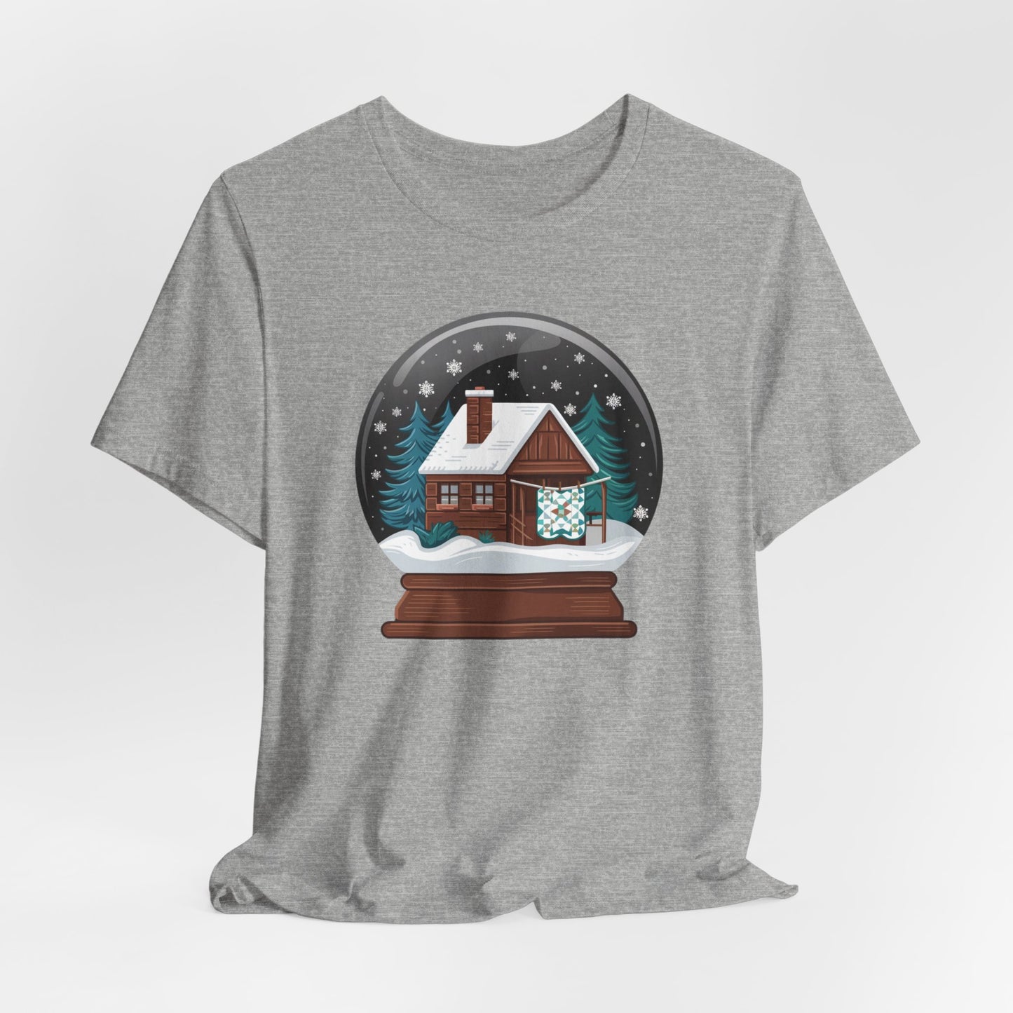 An Athletic Heather christmas quilting t-shirt featuring a cozy log cabin covered in snow, with a quilt hanging outside, set inside a snow globe with swirling snowflakes, blending quilting and holiday warmth.