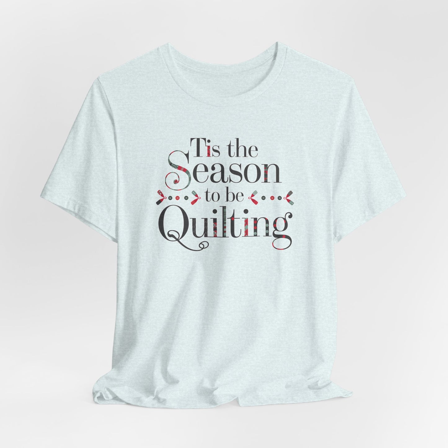 A Heather Ice Blue festive t-shirt featuring the phrase "'Tis the Season to be Quilting," with playful sewing-inspired elements like needles, buttons, and fabric patterns integrated into the typography.