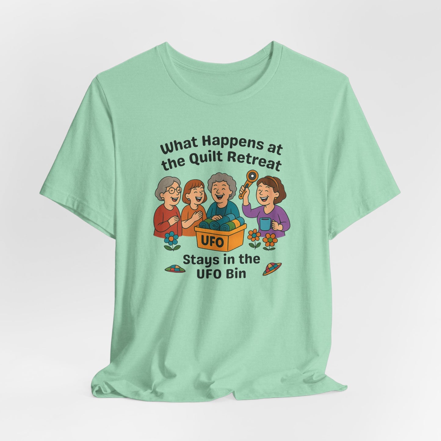 A A Heather Mint t-shirt featuring a cartoon design of four laughing women at a quilt retreat with a bin labeled “UFO” full of fabric. Surrounded by patchwork flying saucers and flowers. Text reads “What happens at the quilt retreat stays in the UFO bin.”