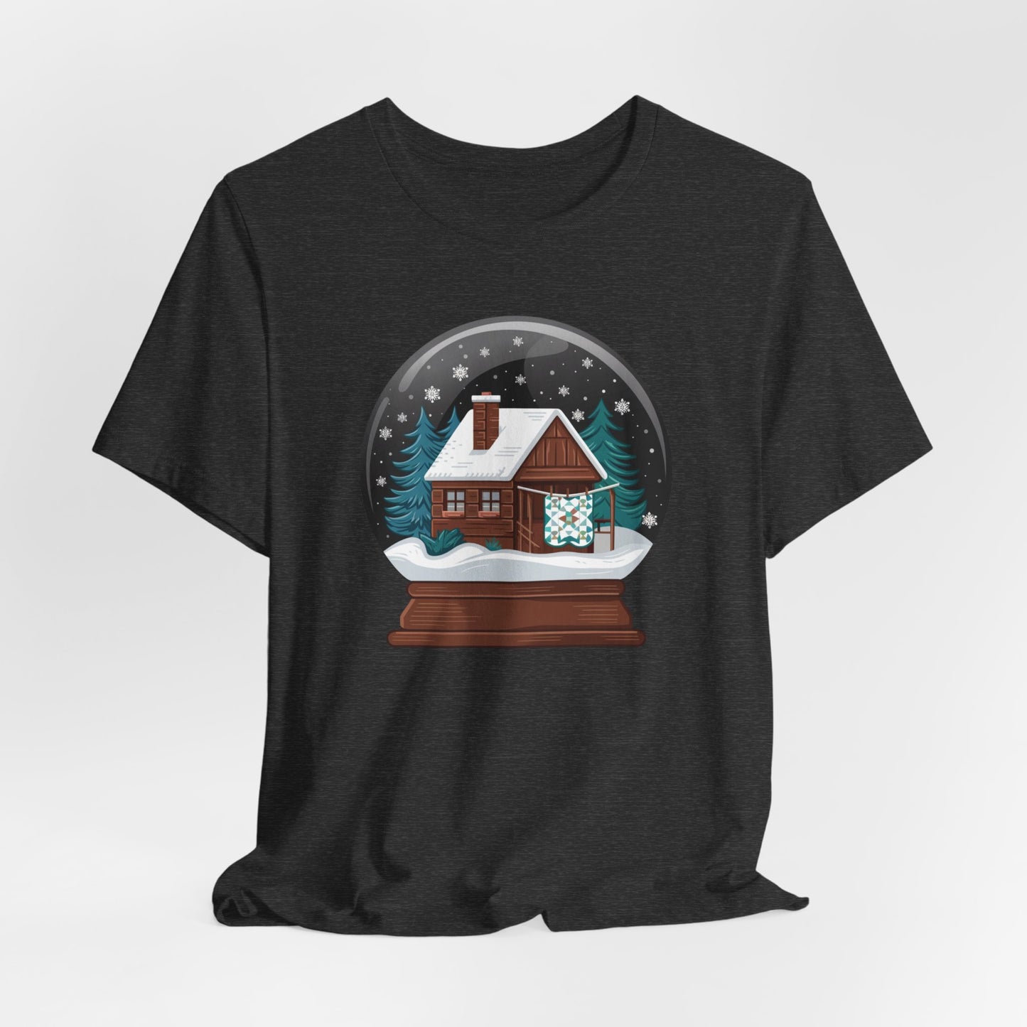 A Dark Grey Heather christmas quilting t-shirt featuring a cozy log cabin covered in snow, with a quilt hanging outside, set inside a snow globe with swirling snowflakes, blending quilting and holiday warmth.