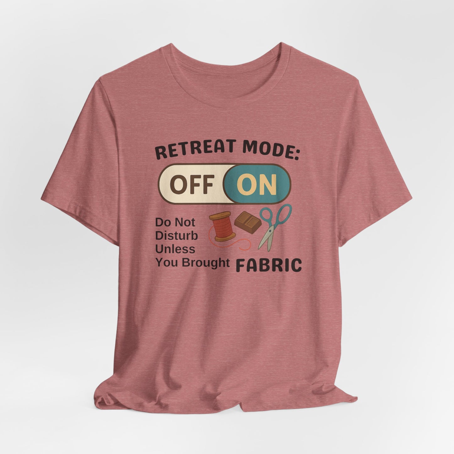 A A Heather Mauve t-shirt with a toggle switch that says "Retreat Mode: OFF/ON" surrounded by a spool of thread, chocolate, and scissors. Text below reads "Do not disturb unless you brought fabric." Fun gift for quilting retreats.