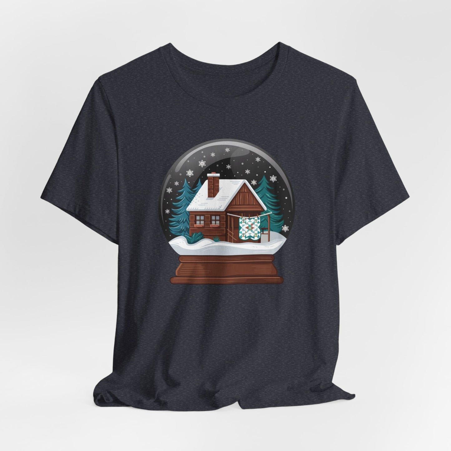 A Heather Navy christmas quilting t-shirt featuring a cozy log cabin covered in snow, with a quilt hanging outside, set inside a snow globe with swirling snowflakes, blending quilting and holiday warmth.