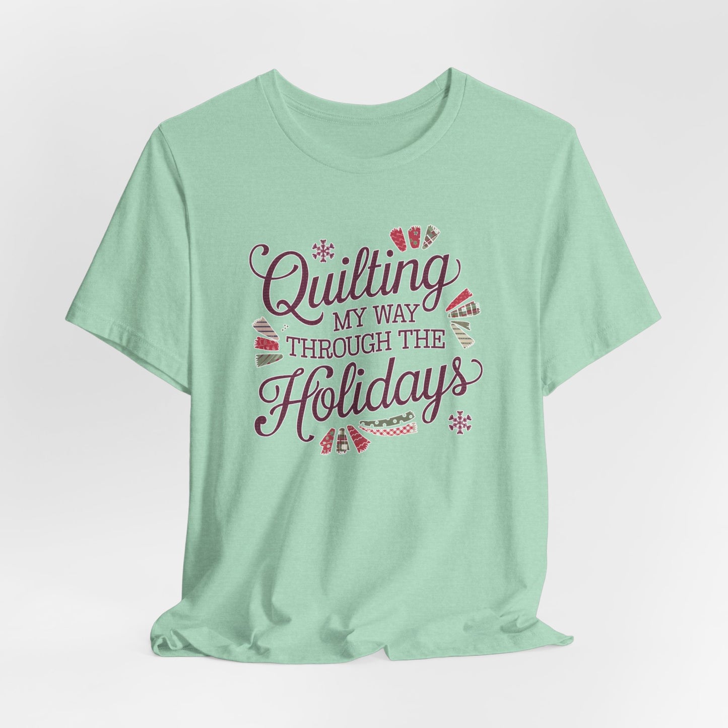 A Heather Mint playful holiday-themed t-shirt with the phrase "Quilting My Way Through the Holidays" surrounded by quilting-inspired decorations like fabric swatches and snowflakes.