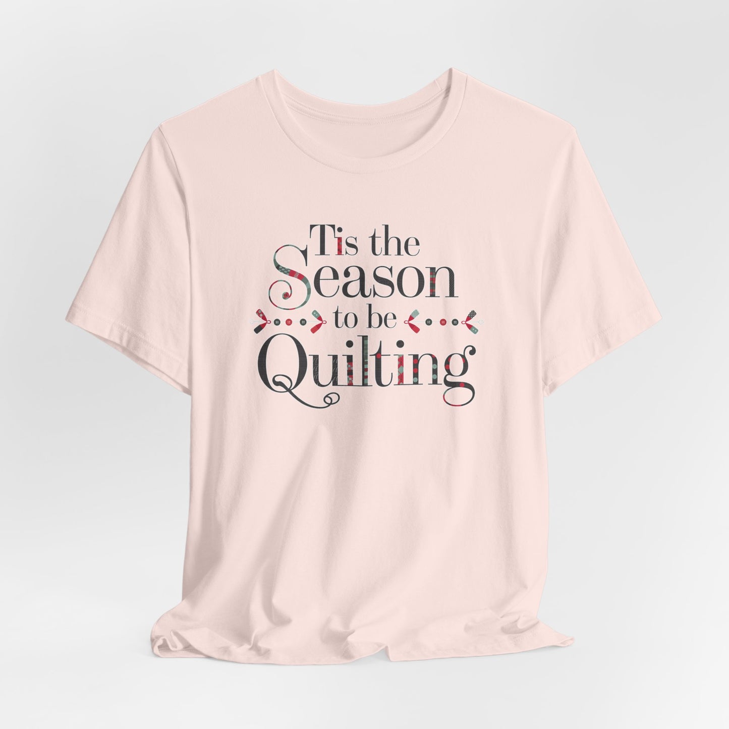 A Soft Pink festive t-shirt featuring the phrase "'Tis the Season to be Quilting," with playful sewing-inspired elements like needles, buttons, and fabric patterns integrated into the typography.