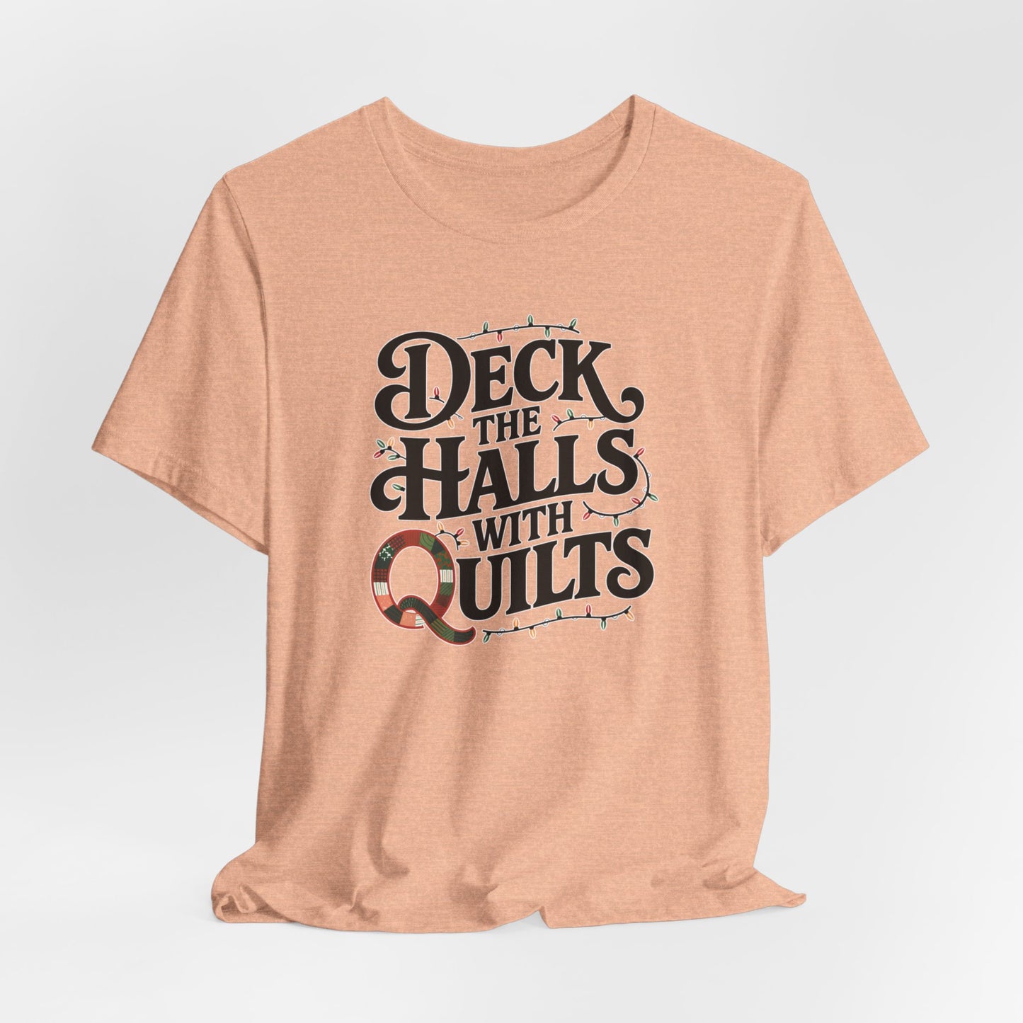 A Heather Peach christmas quilting t-shirt featuring the phrase "Deck the Halls with Quilts," adorned with holiday lights and quilting-inspired elements, including a quilted 'Q' for a humorous touch.