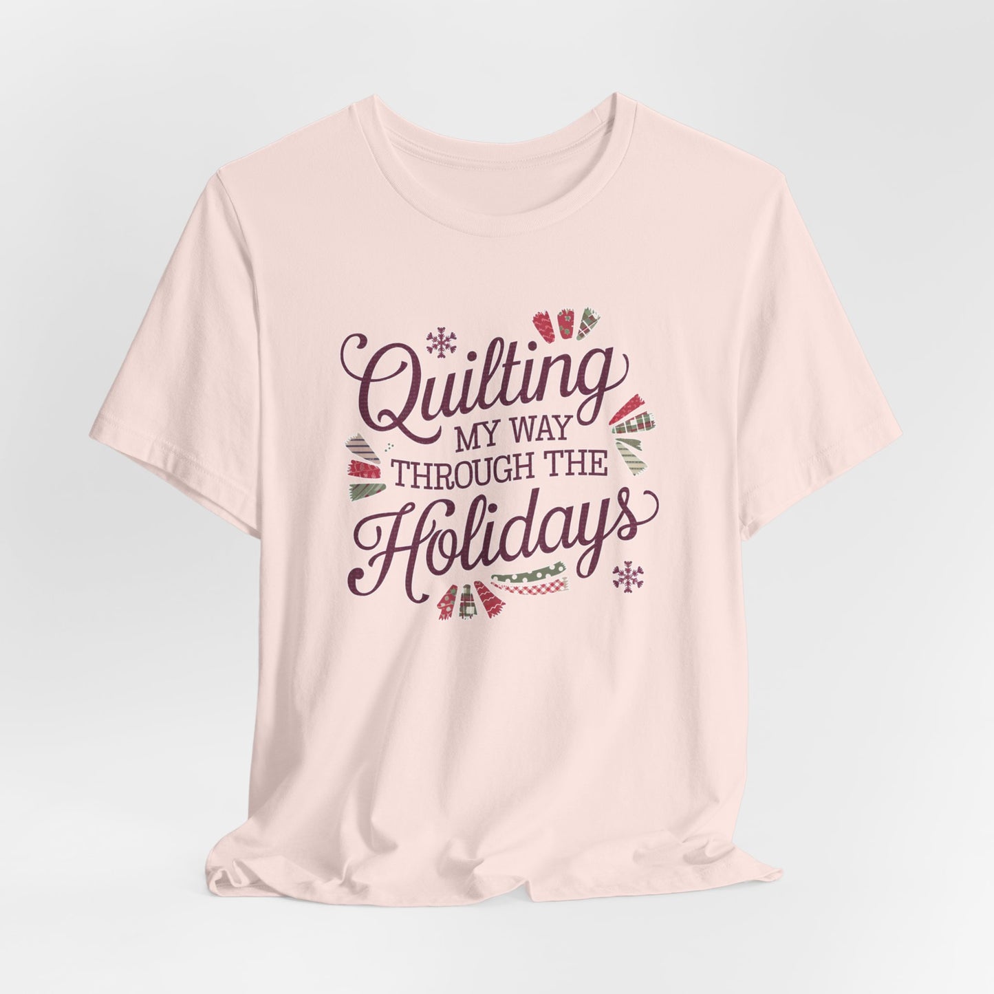 A Soft Pink playful holiday-themed t-shirt with the phrase "Quilting My Way Through the Holidays" surrounded by quilting-inspired decorations like fabric swatches and snowflakes.