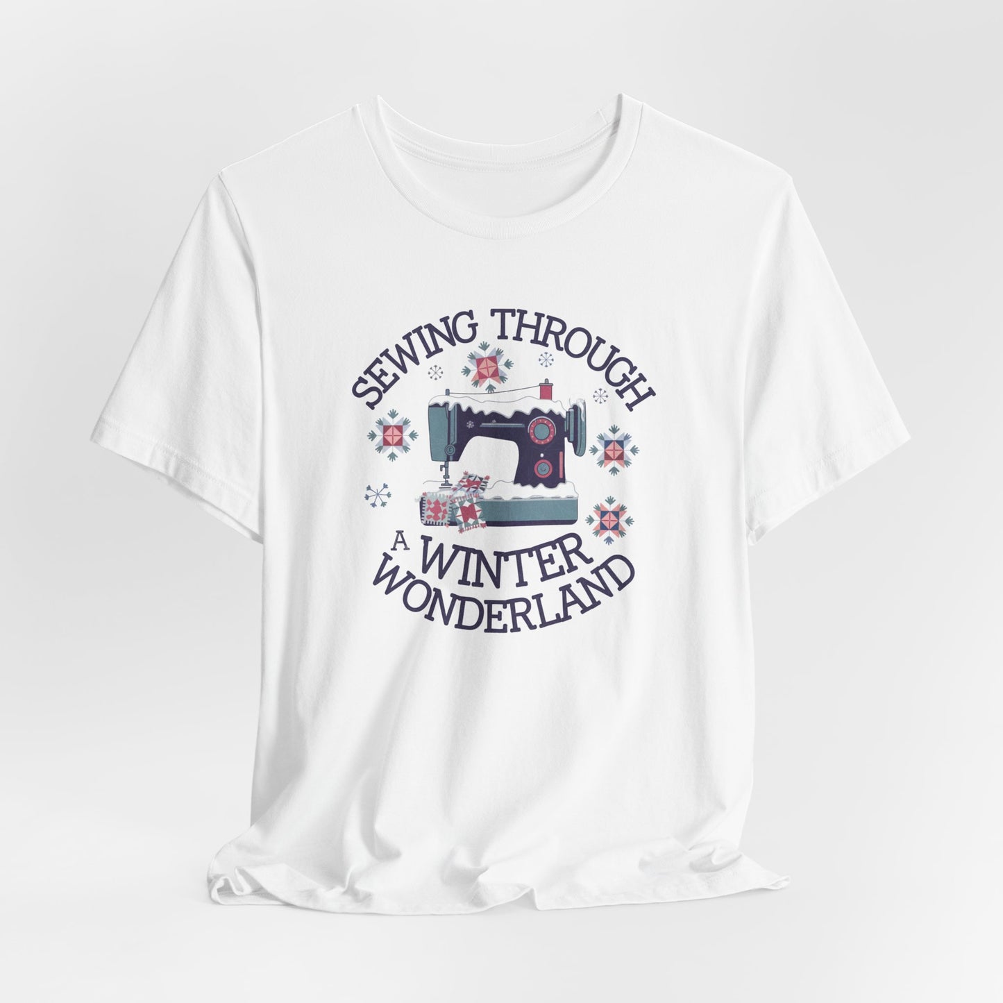 A White winter quilting t-shirt featuring a sewing machine covered in snow, surrounded by quilted snowflakes, with the phrase "Sewing Through a Winter Wonderland" above and below the image.