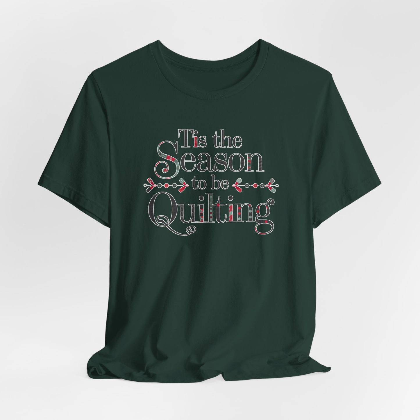 A Forest festive t-shirt featuring the phrase "'Tis the Season to be Quilting," with playful sewing-inspired elements like needles, buttons, and fabric patterns integrated into the typography.
