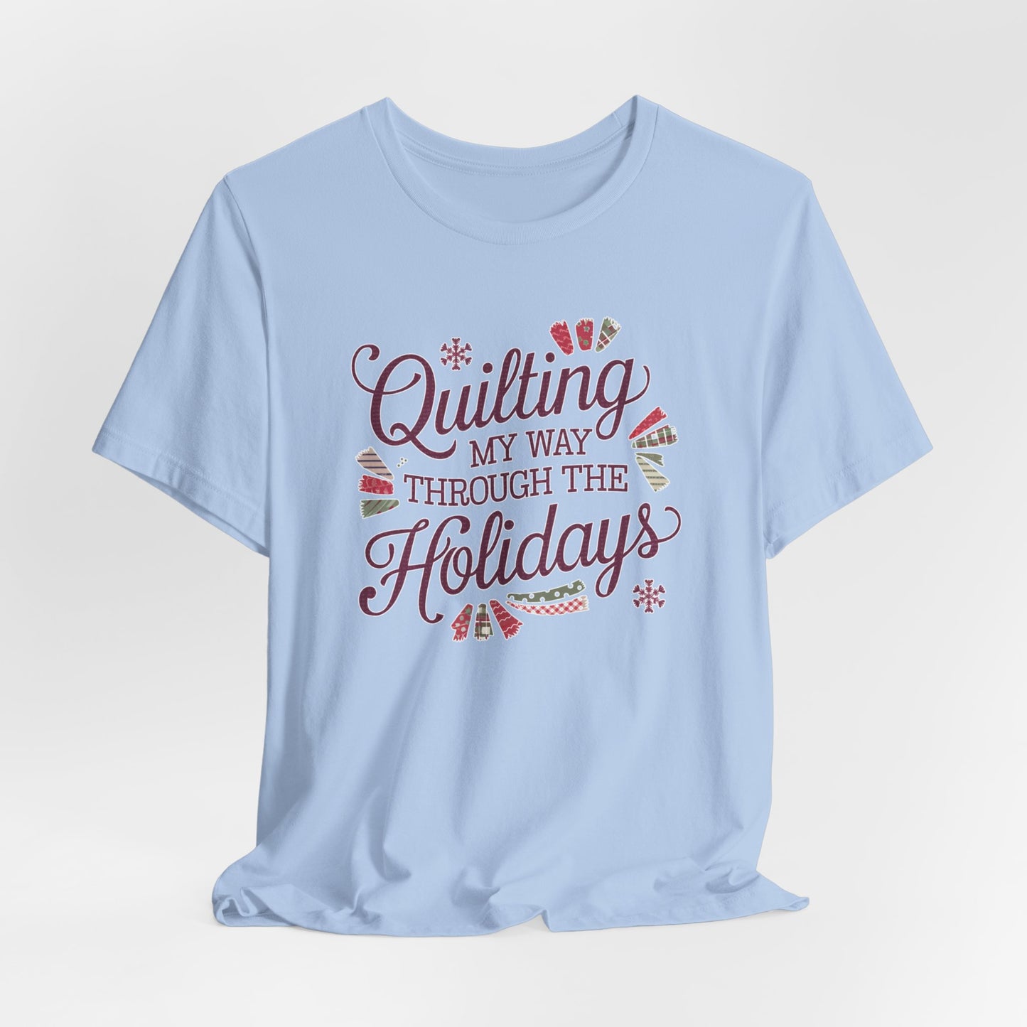 A Baby Blue playful holiday-themed t-shirt with the phrase "Quilting My Way Through the Holidays" surrounded by quilting-inspired decorations like fabric swatches and snowflakes.