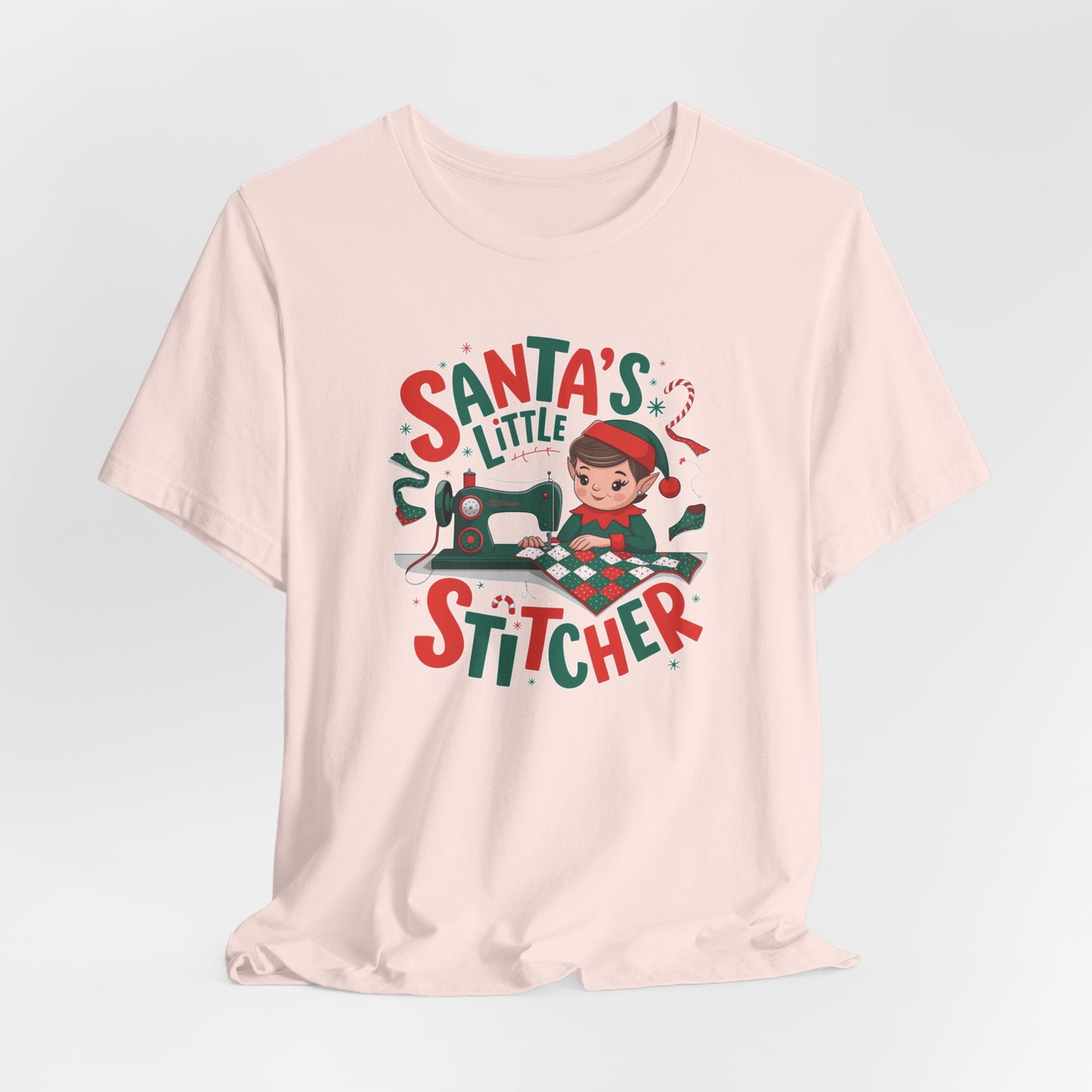 A Soft Pink christmas quilting t-shirt featuring an adorable elf is sewing a festive patchwork quilt on a sewing machine, surrounded by holiday-themed decorations, with the phrase "Santa's Little Stitcher" above and below the design.