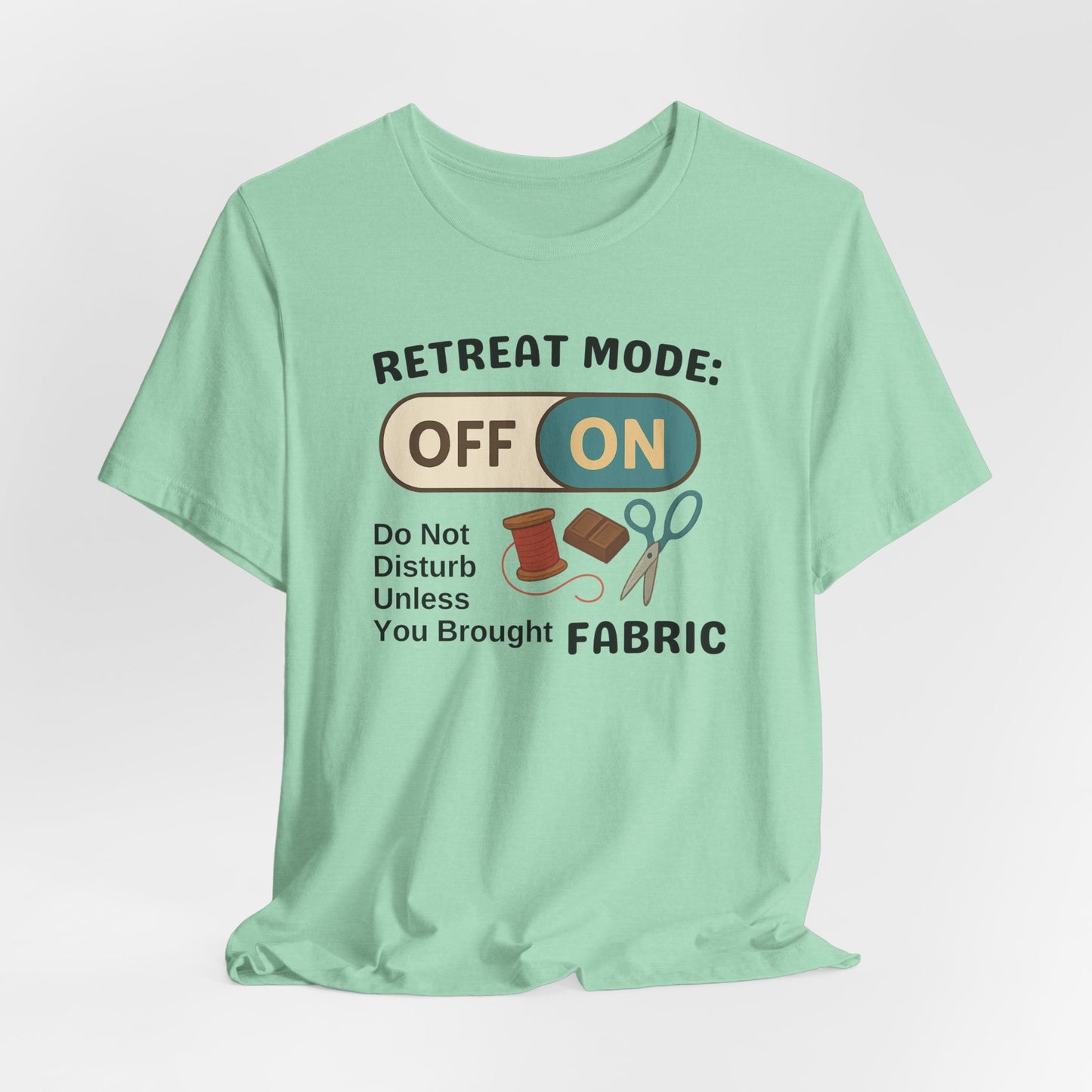 A A Heather Mint t-shirt with a toggle switch that says "Retreat Mode: OFF/ON" surrounded by a spool of thread, chocolate, and scissors. Text below reads "Do not disturb unless you brought fabric." Fun gift for quilting retreats.
