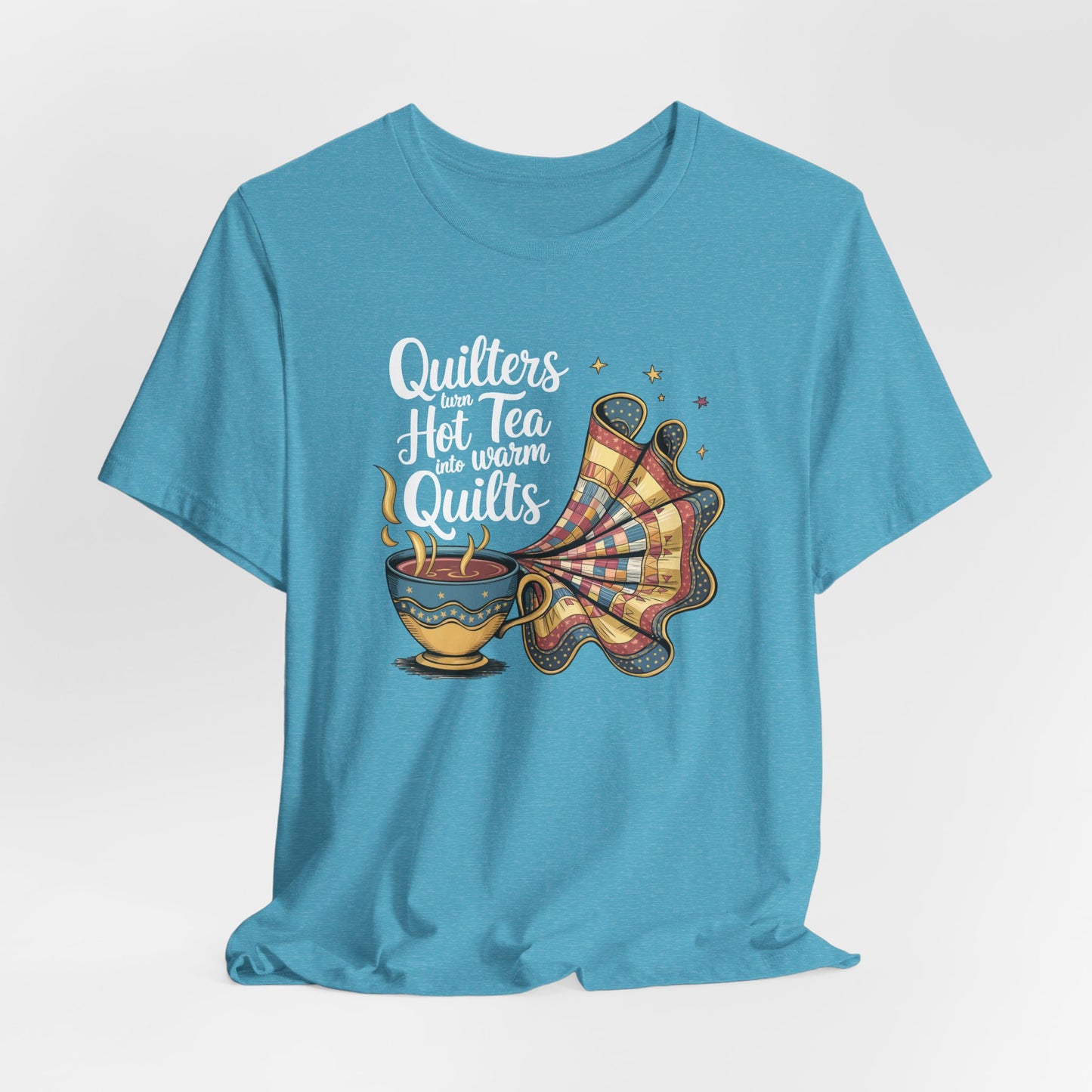 A Heather Aqua t-shirt with a whimsical design of a teacup transforming into a patchwork quilt, accompanied by the phrase 'Quilters turn hot tea into warm quilts.' Ideal for quilters and tea lovers