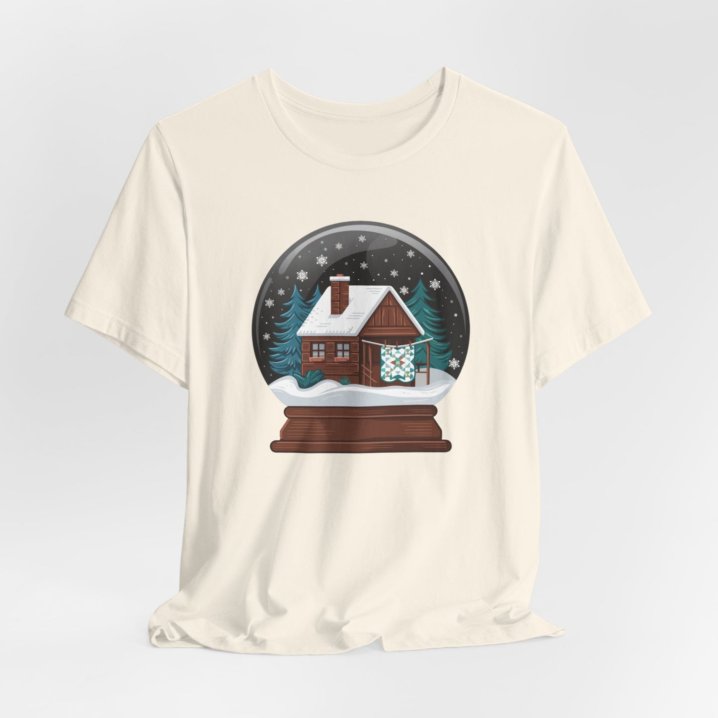 A Natural christmas quilting t-shirt featuring a cozy log cabin covered in snow, with a quilt hanging outside, set inside a snow globe with swirling snowflakes, blending quilting and holiday warmth.