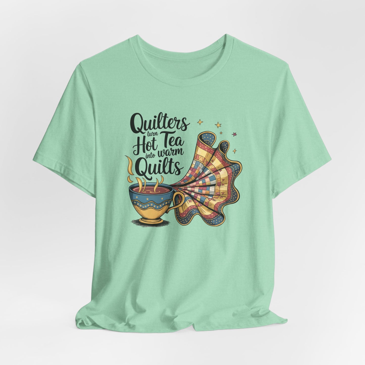 A Heather Mint t-shirt with a whimsical design of a teacup transforming into a patchwork quilt, accompanied by the phrase 'Quilters turn hot tea into warm quilts.' Ideal for quilters and tea lovers
