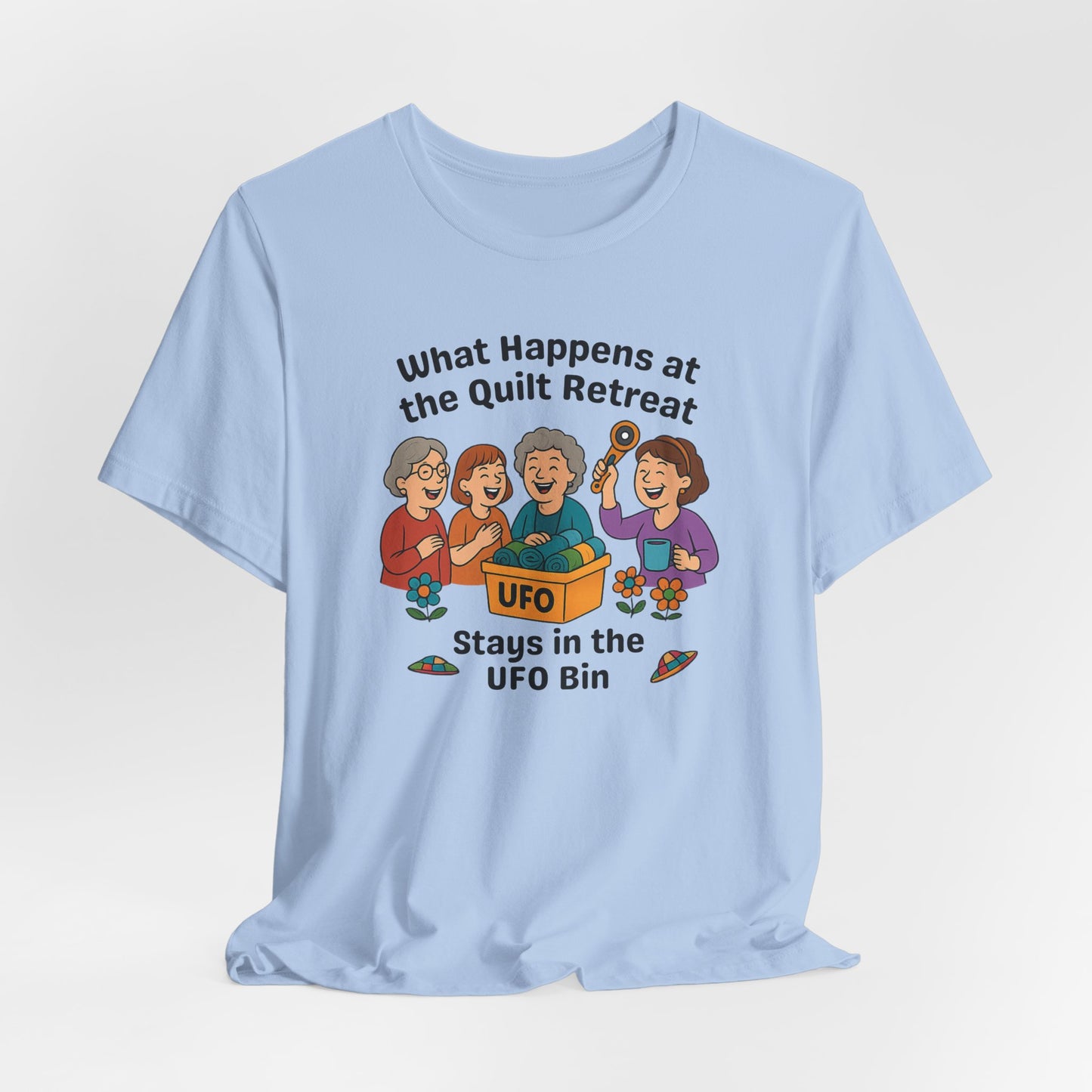 A A Baby Blue t-shirt featuring a cartoon design of four laughing women at a quilt retreat with a bin labeled “UFO” full of fabric. Surrounded by patchwork flying saucers and flowers. Text reads “What happens at the quilt retreat stays in the UFO bin.”