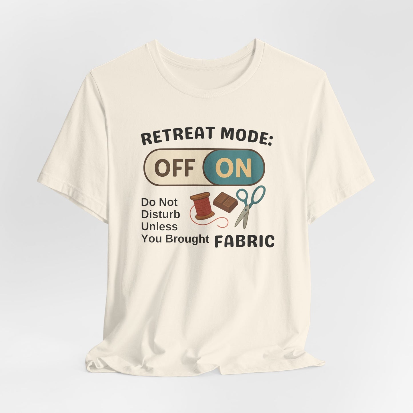 A A Natural t-shirt with a toggle switch that says "Retreat Mode: OFF/ON" surrounded by a spool of thread, chocolate, and scissors. Text below reads "Do not disturb unless you brought fabric." Fun gift for quilting retreats.