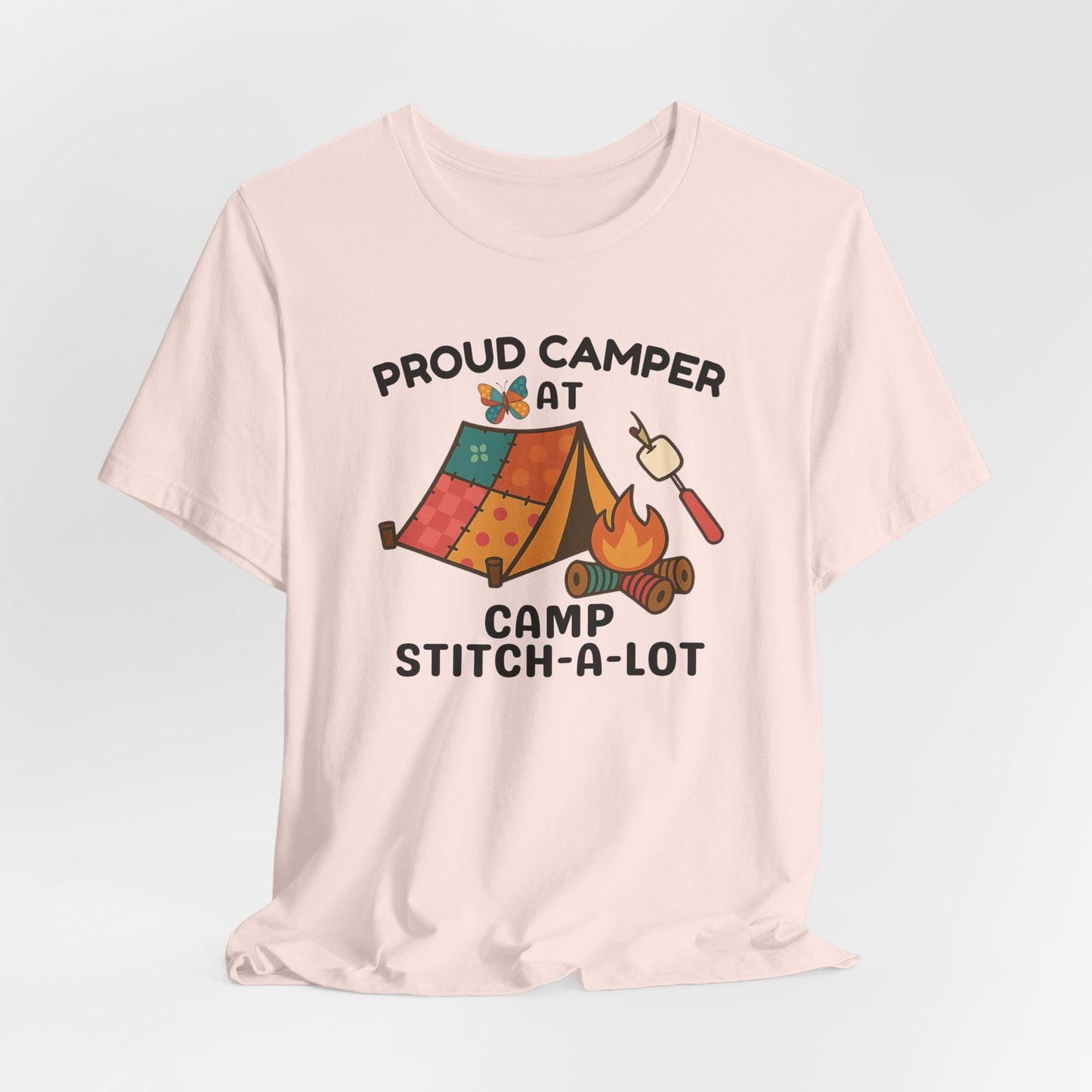 A A Soft Pink t-shirt featuring a colorful design with a patchwork tent, thread spool campfire, marshmallow on a seam ripper stick, and a butterfly. Text reads "Proud Camper at Camp Stitch-a-Lot." Fun gift for quilting retreats.