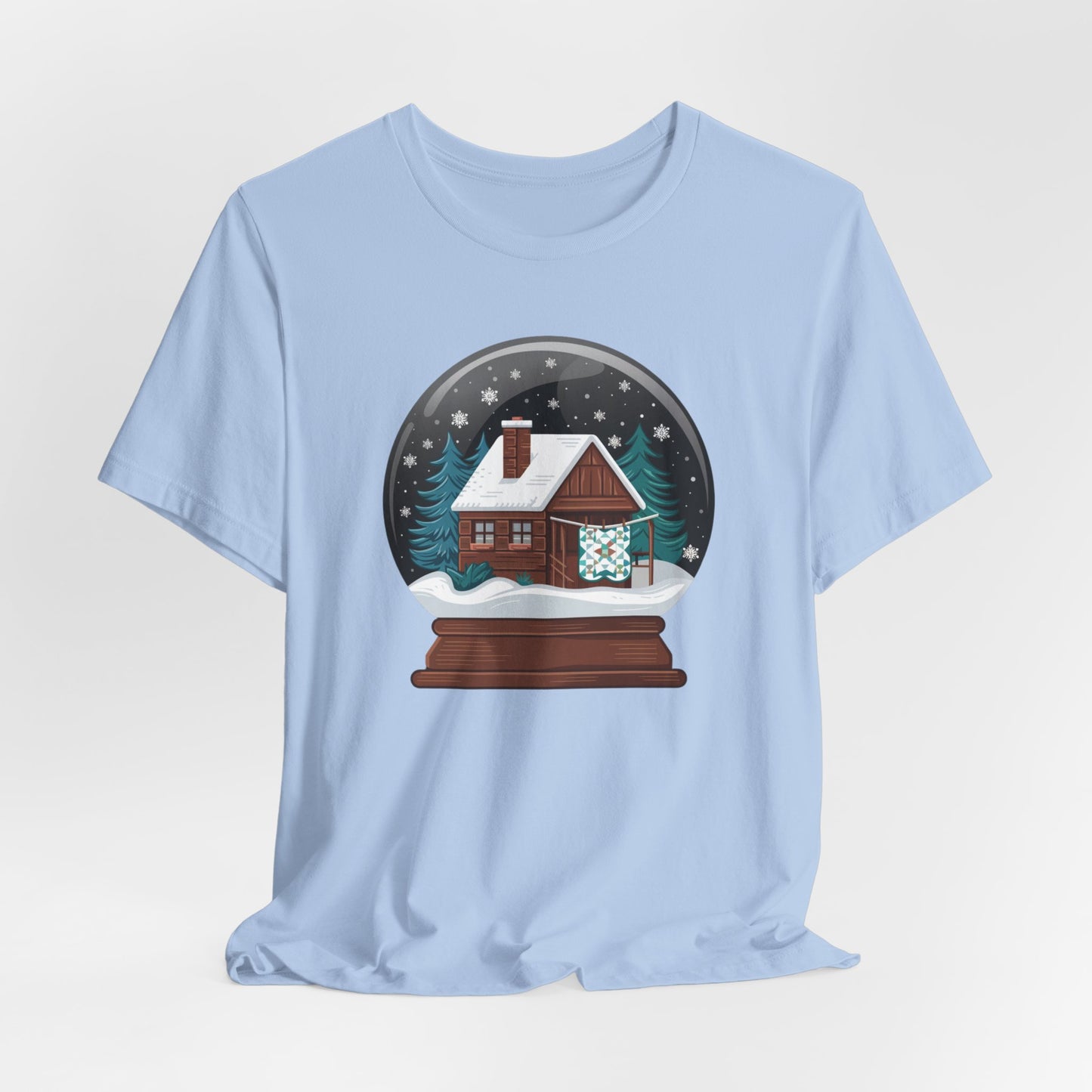 A Baby Blue christmas quilting t-shirt featuring a cozy log cabin covered in snow, with a quilt hanging outside, set inside a snow globe with swirling snowflakes, blending quilting and holiday warmth.