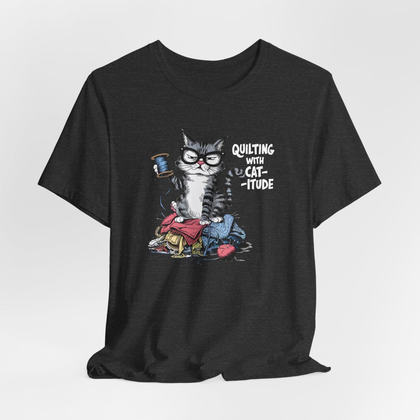 A Dark Grey Heather t-shirt featuring a sassy cartoon cat sitting on a pile of colorful fabric, holding a spool of thread, with the phrase 'Quilting with Cat-itude,' perfect for quilters and cat lovers.