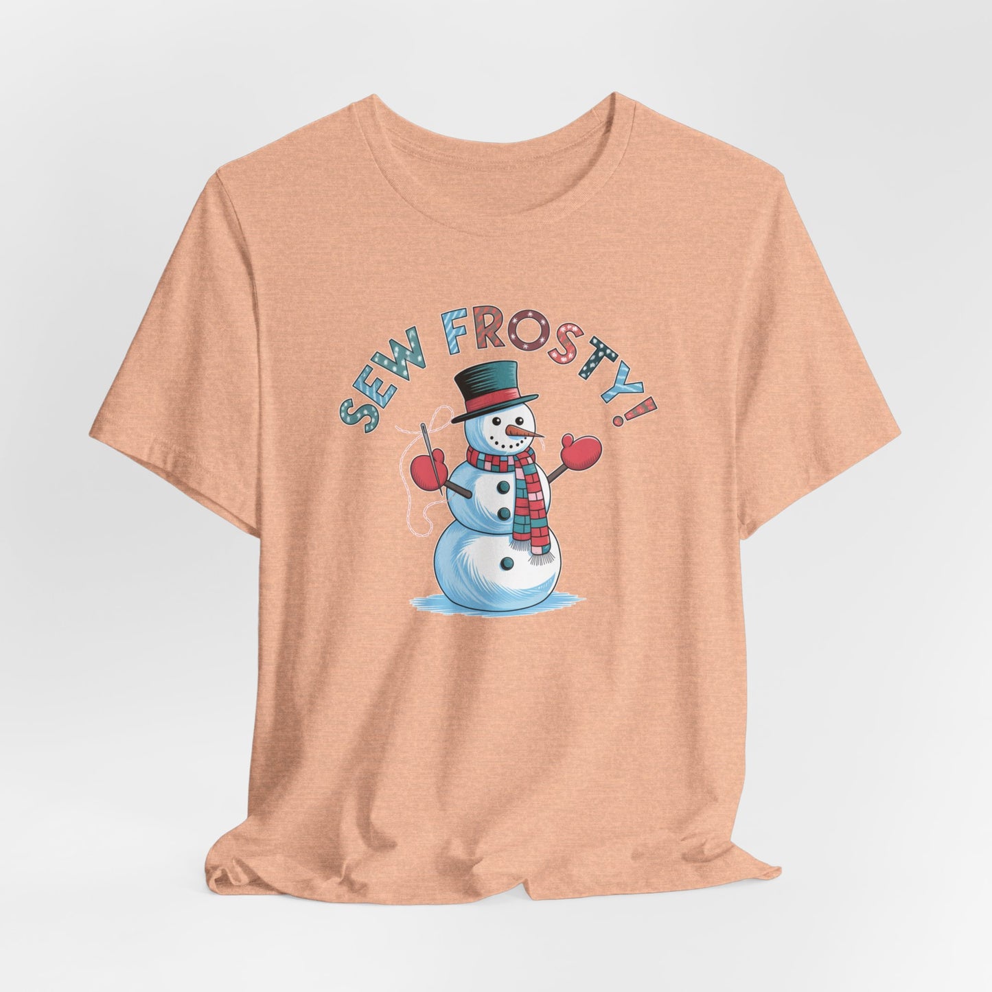 A Heather Peach christmas quilting t-shirt featuring cheerful snowman design holding a sewing needle, with the phrase "Sew Frosty" in playful, winter-themed lettering. The snowman is wearing a scarf and mittens, ready for holiday sewing fun.
