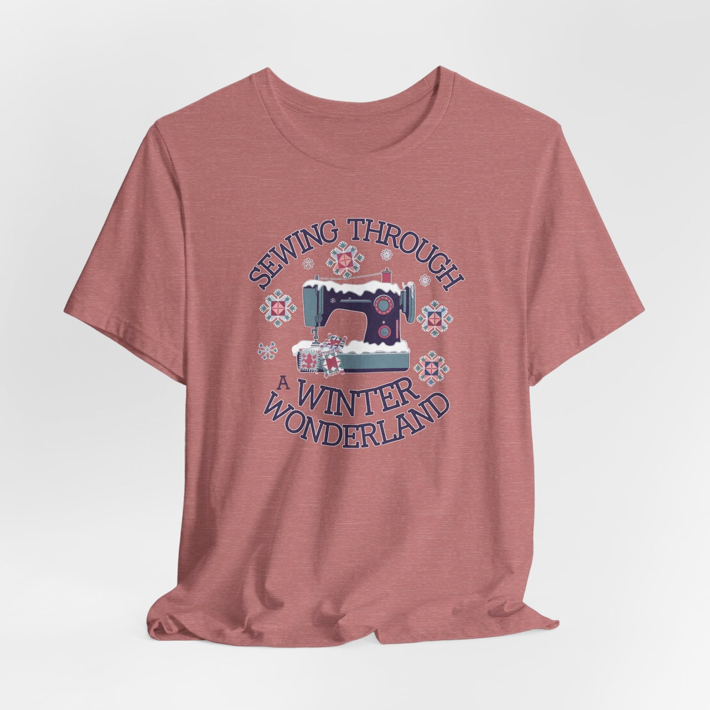 A Heather Mauve winter quilting t-shirt featuring a sewing machine covered in snow, surrounded by quilted snowflakes, with the phrase "Sewing Through a Winter Wonderland" above and below the image.