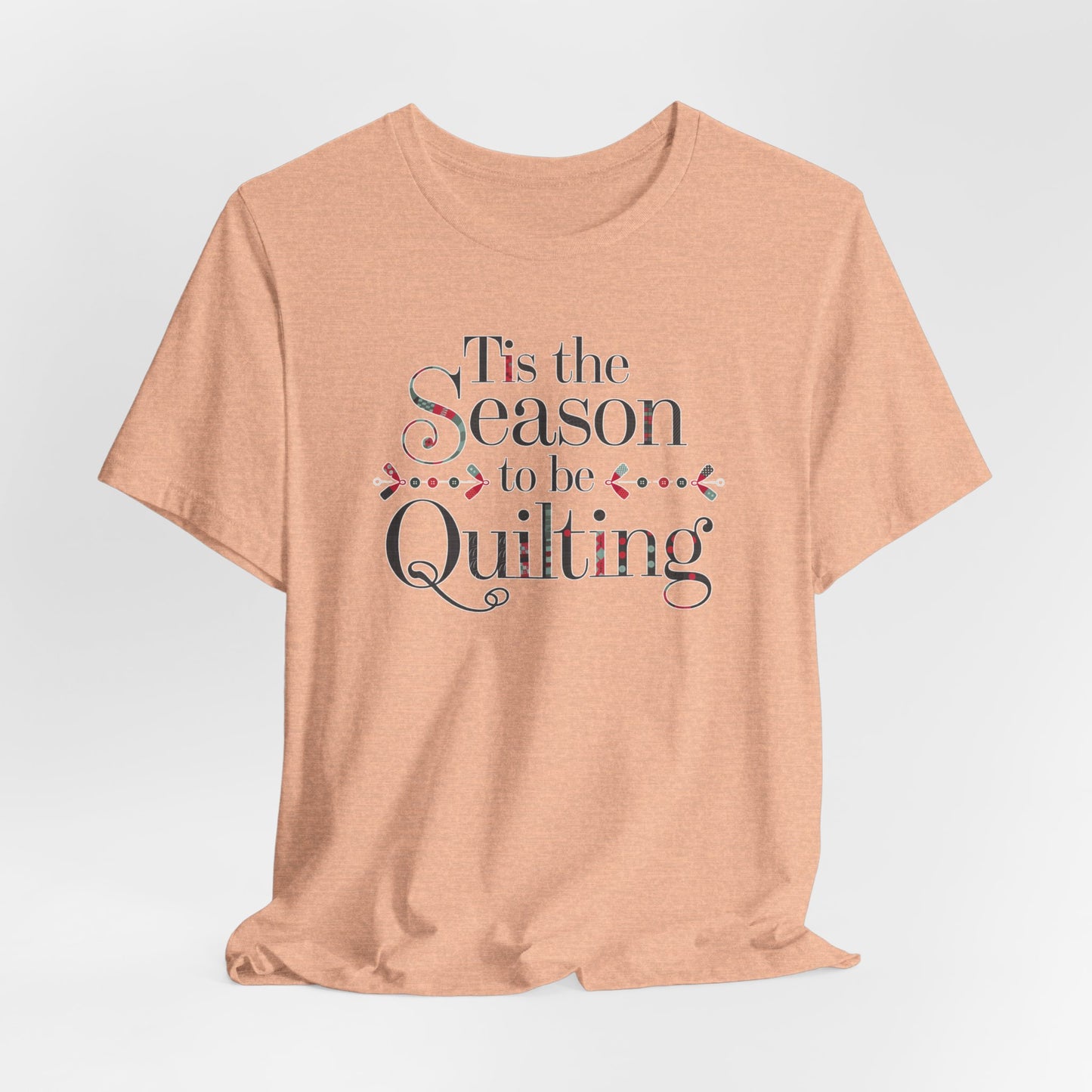 A Heather Peach festive t-shirt featuring the phrase "'Tis the Season to be Quilting," with playful sewing-inspired elements like needles, buttons, and fabric patterns integrated into the typography.