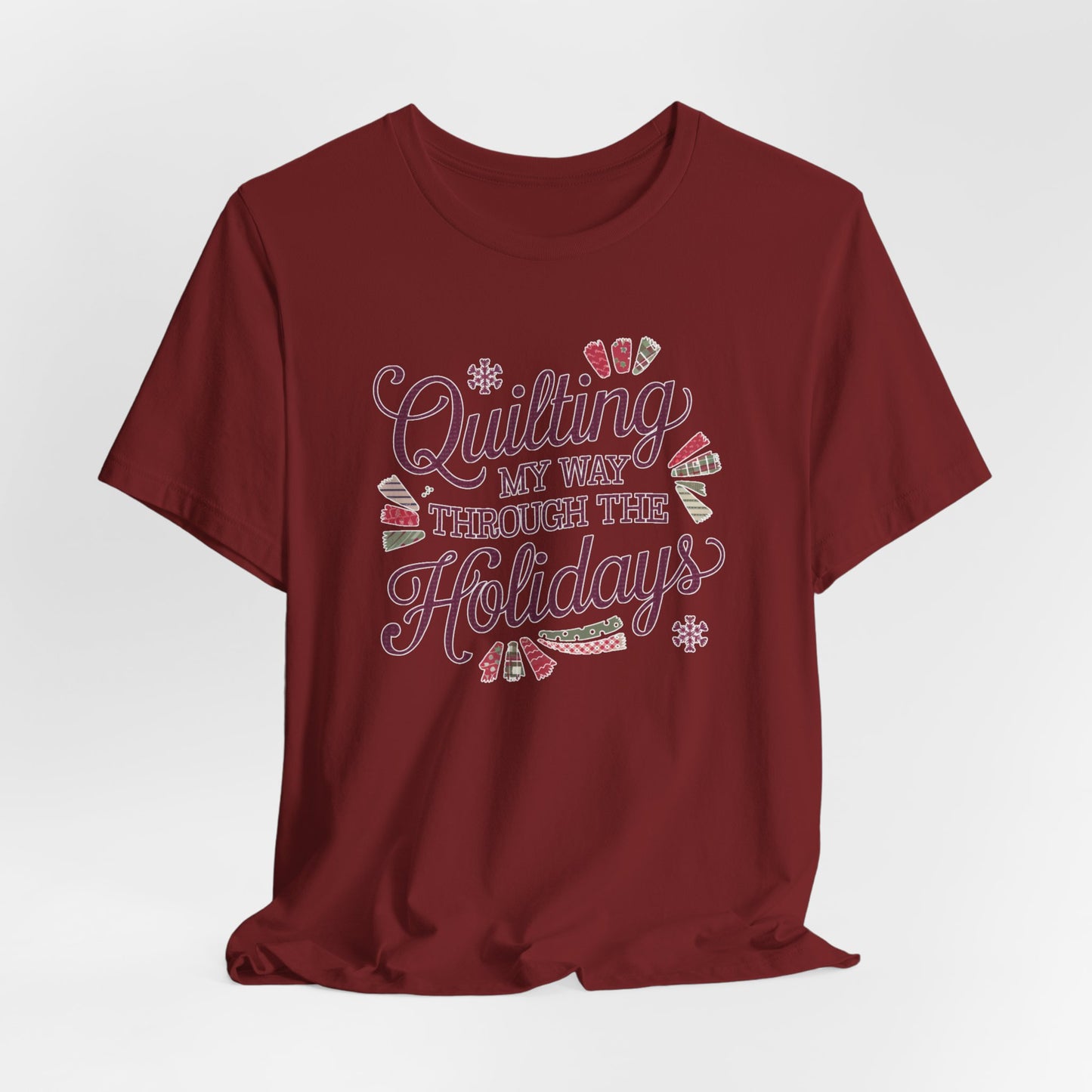 A Cardinal playful holiday-themed t-shirt with the phrase "Quilting My Way Through the Holidays" surrounded by quilting-inspired decorations like fabric swatches and snowflakes.