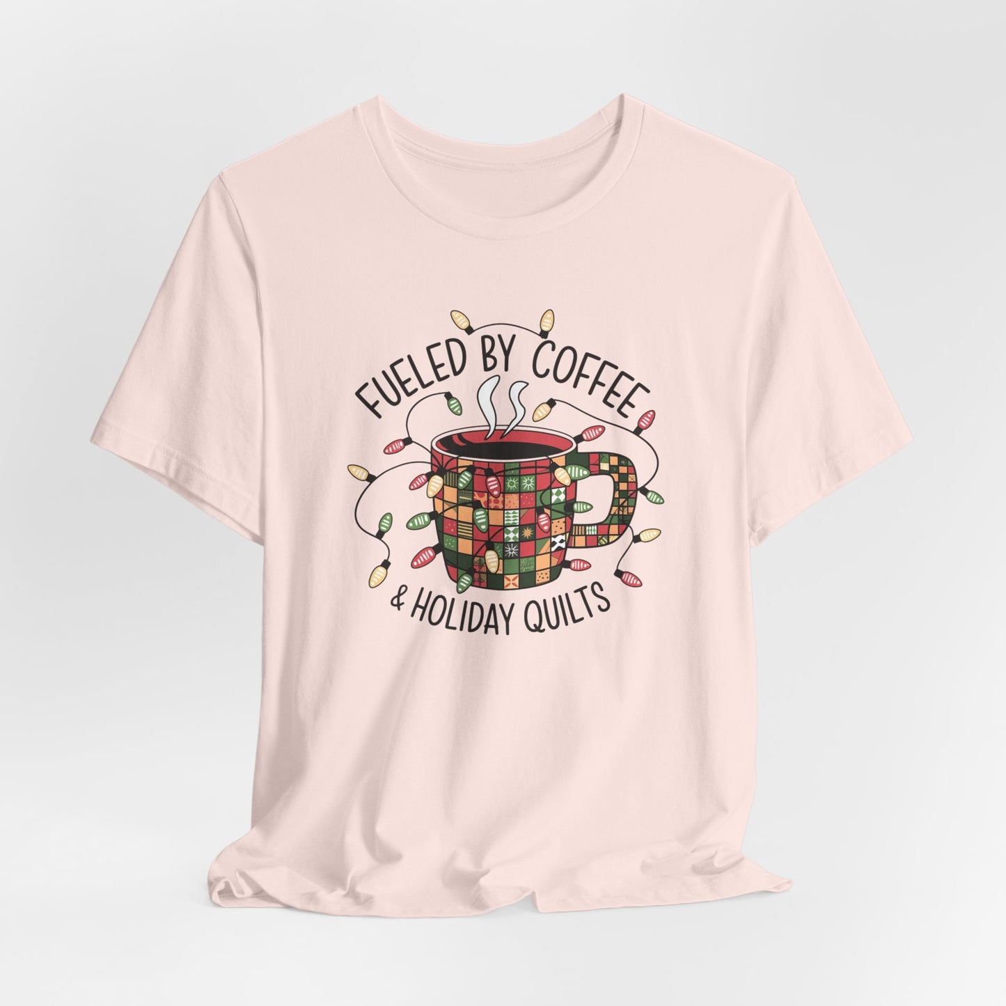 A Soft Pink christmas quilting t-shirt featuring a cozy coffee mug covered in patchwork quilt patterns, wrapped in festive holiday lights, with the phrase "Fueled by Coffee & Holiday Quilts" surrounding the design.