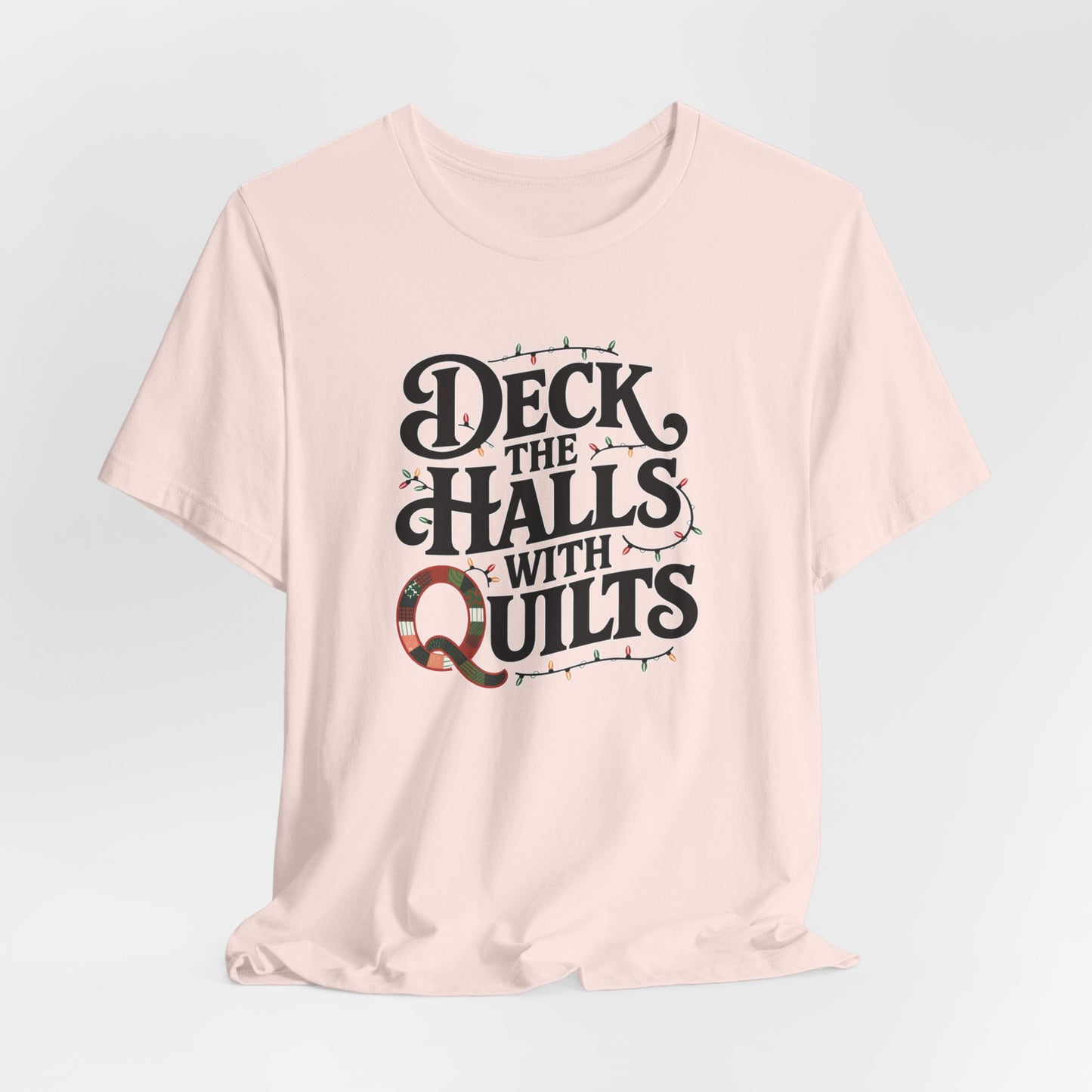 A Soft Pink christmas quilting t-shirt featuring the phrase "Deck the Halls with Quilts," adorned with holiday lights and quilting-inspired elements, including a quilted 'Q' for a humorous touch.