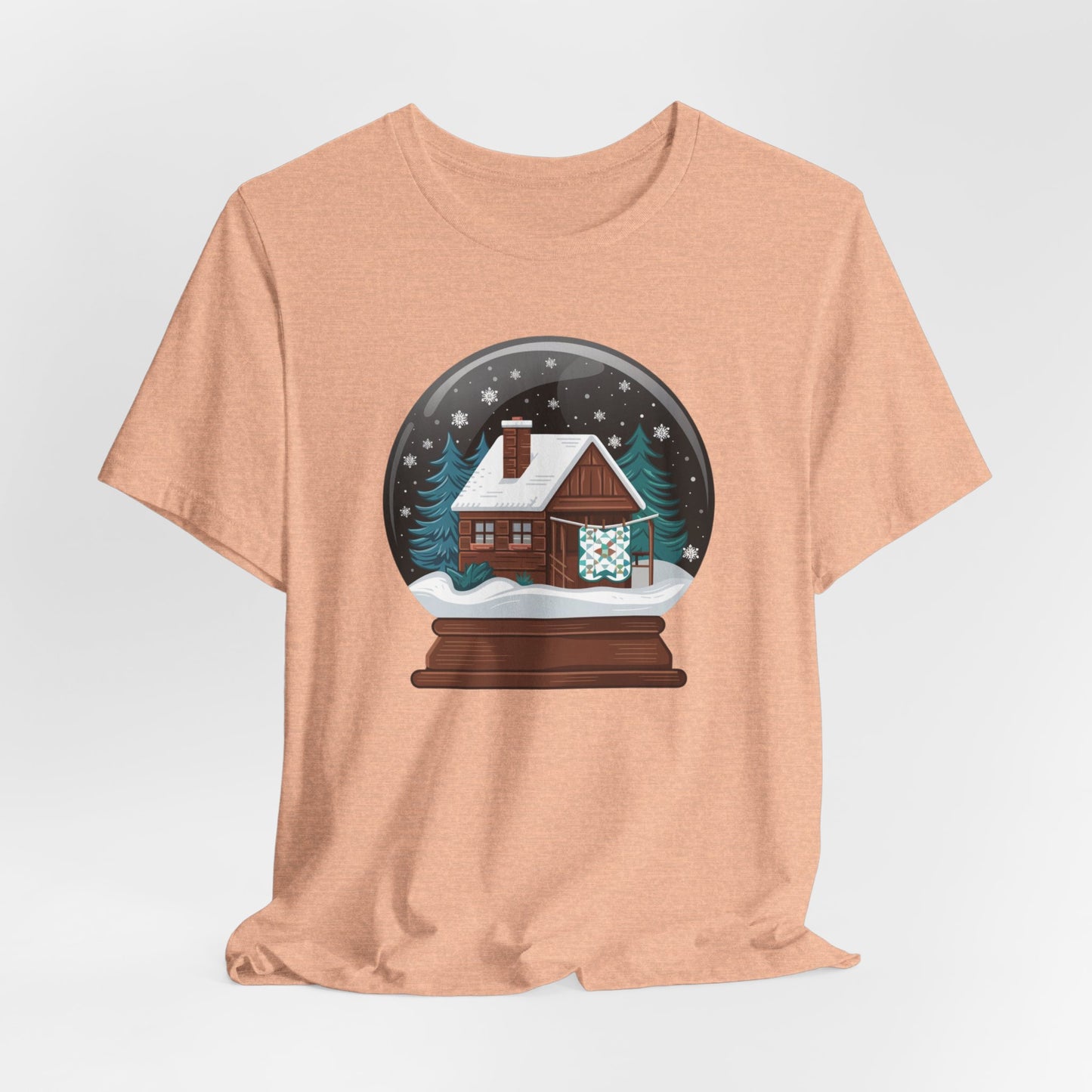 A Heather Peach christmas quilting t-shirt featuring a cozy log cabin covered in snow, with a quilt hanging outside, set inside a snow globe with swirling snowflakes, blending quilting and holiday warmth.