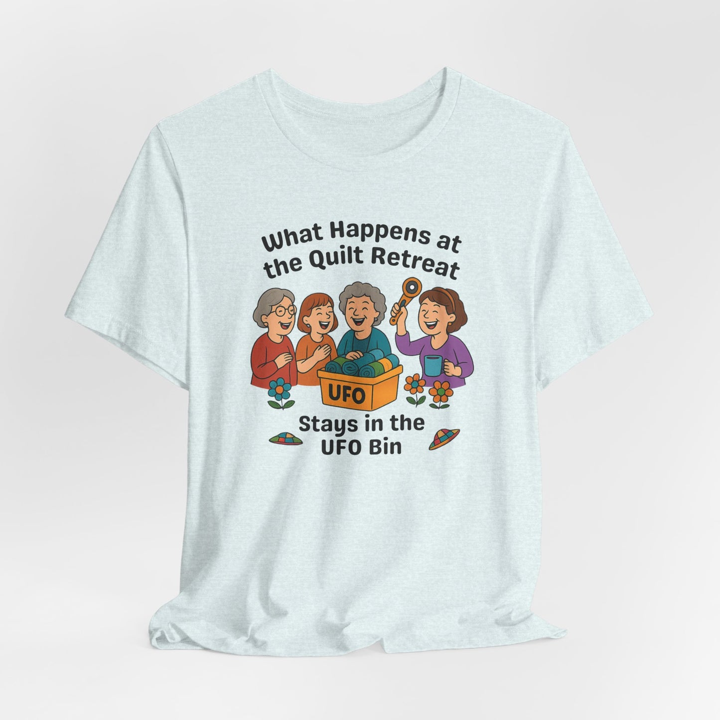 A A Heather Ice Blue t-shirt featuring a cartoon design of four laughing women at a quilt retreat with a bin labeled “UFO” full of fabric. Surrounded by patchwork flying saucers and flowers. Text reads “What happens at the quilt retreat stays in the UFO bin.”