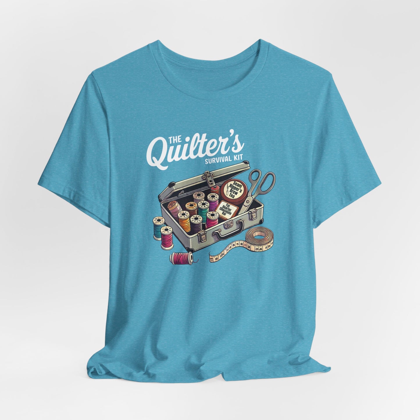 A Heather Aqua t-shirt featuring a whimsical illustration of a quilter’s survival kit with colorful thread spools, scissors, and a tape measure, perfect for sewing and quilting enthusiasts.