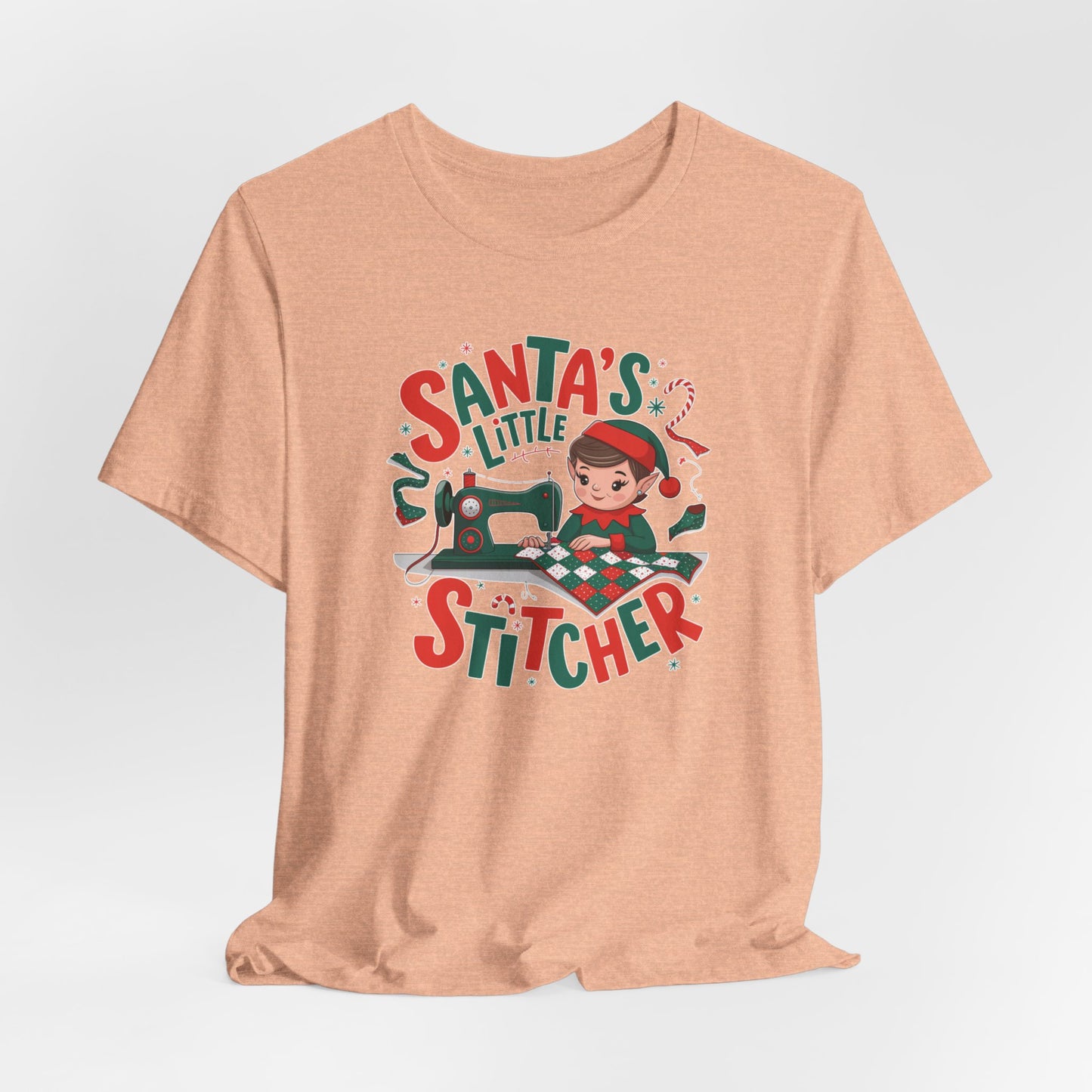 A Heather Peach christmas quilting t-shirt featuring an adorable elf is sewing a festive patchwork quilt on a sewing machine, surrounded by holiday-themed decorations, with the phrase "Santa's Little Stitcher" above and below the design.