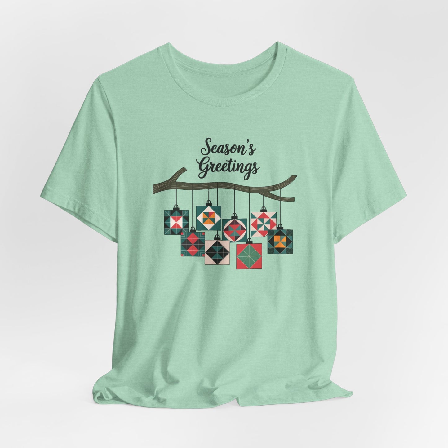 A Heather Mint christmas quilting t-shirt featuring quilt block ornaments hanging from a branch, featuring the phrase "Season's Greetings" above. The ornaments showcase various quilting patterns to highlight the holiday quilting theme.