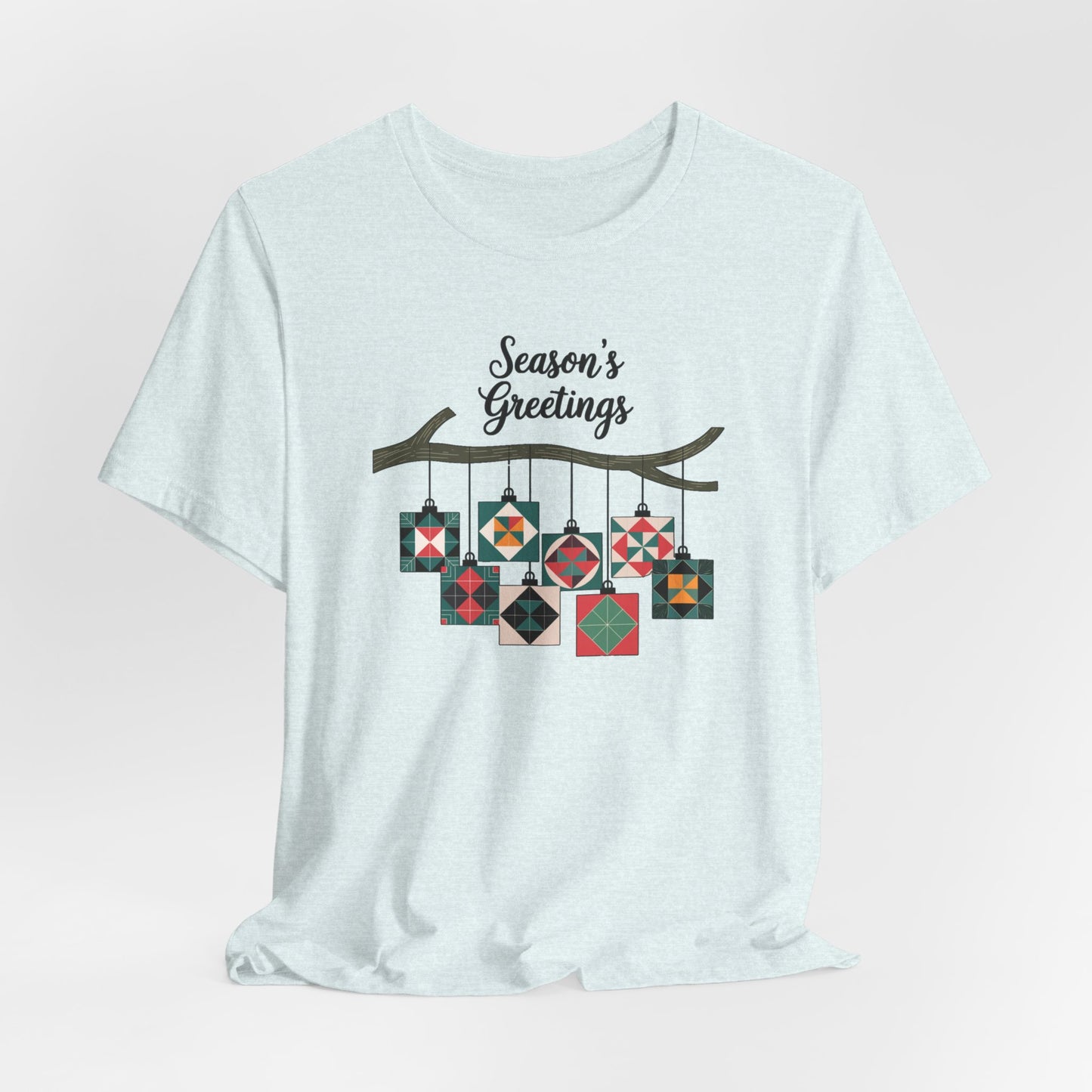 A Heather Ice Blue christmas quilting t-shirt featuring quilt block ornaments hanging from a branch, featuring the phrase "Season's Greetings" above. The ornaments showcase various quilting patterns to highlight the holiday quilting theme.