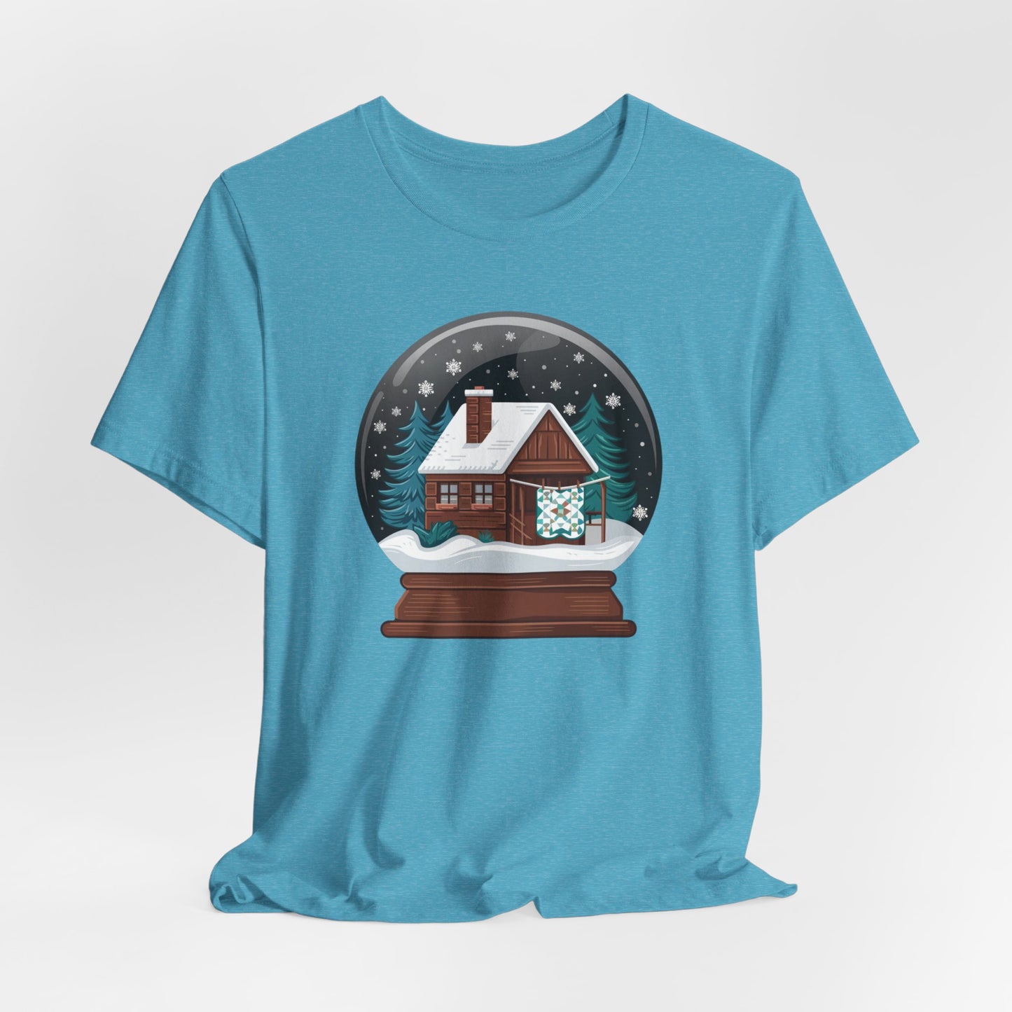 A Heather Aqua christmas quilting t-shirt featuring a cozy log cabin covered in snow, with a quilt hanging outside, set inside a snow globe with swirling snowflakes, blending quilting and holiday warmth.