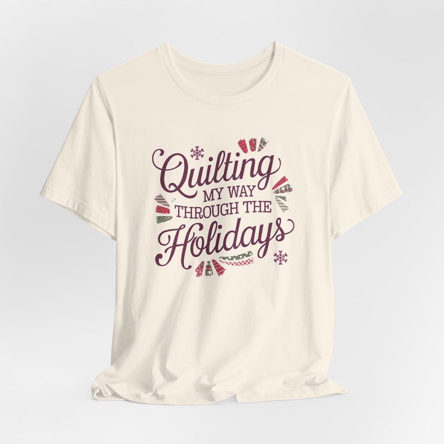 A Natural playful holiday-themed t-shirt with the phrase "Quilting My Way Through the Holidays" surrounded by quilting-inspired decorations like fabric swatches and snowflakes.