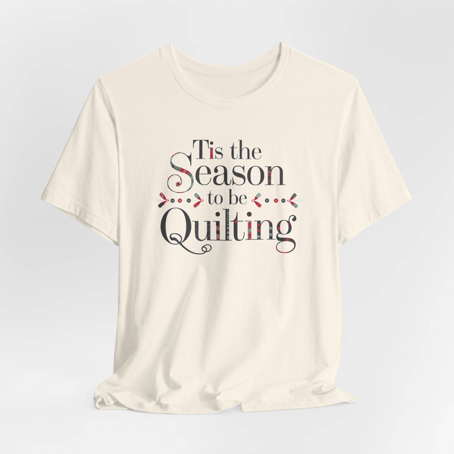 A Natural festive t-shirt featuring the phrase "'Tis the Season to be Quilting," with playful sewing-inspired elements like needles, buttons, and fabric patterns integrated into the typography.