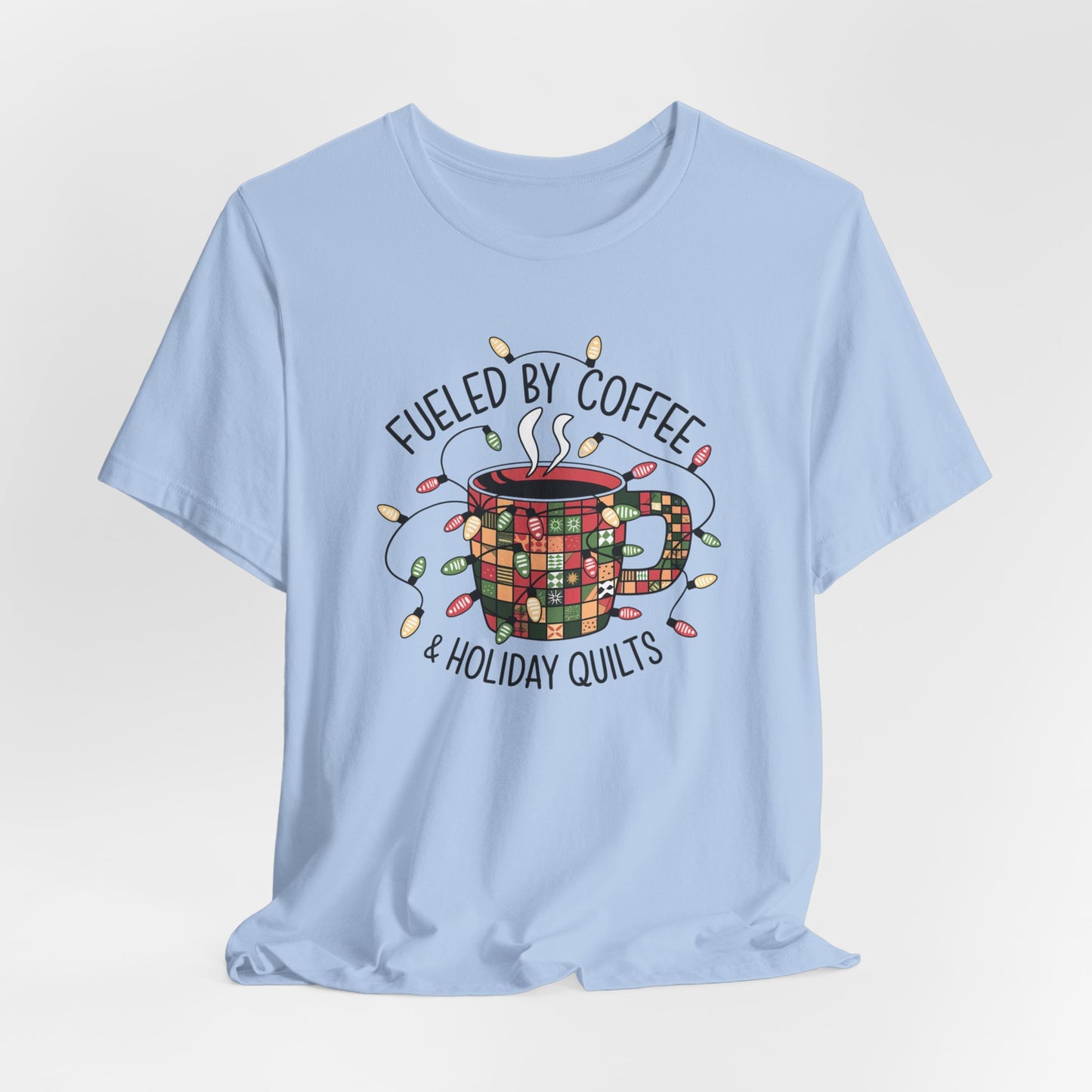 A Baby Blue christmas quilting t-shirt featuring a cozy coffee mug covered in patchwork quilt patterns, wrapped in festive holiday lights, with the phrase "Fueled by Coffee & Holiday Quilts" surrounding the design.