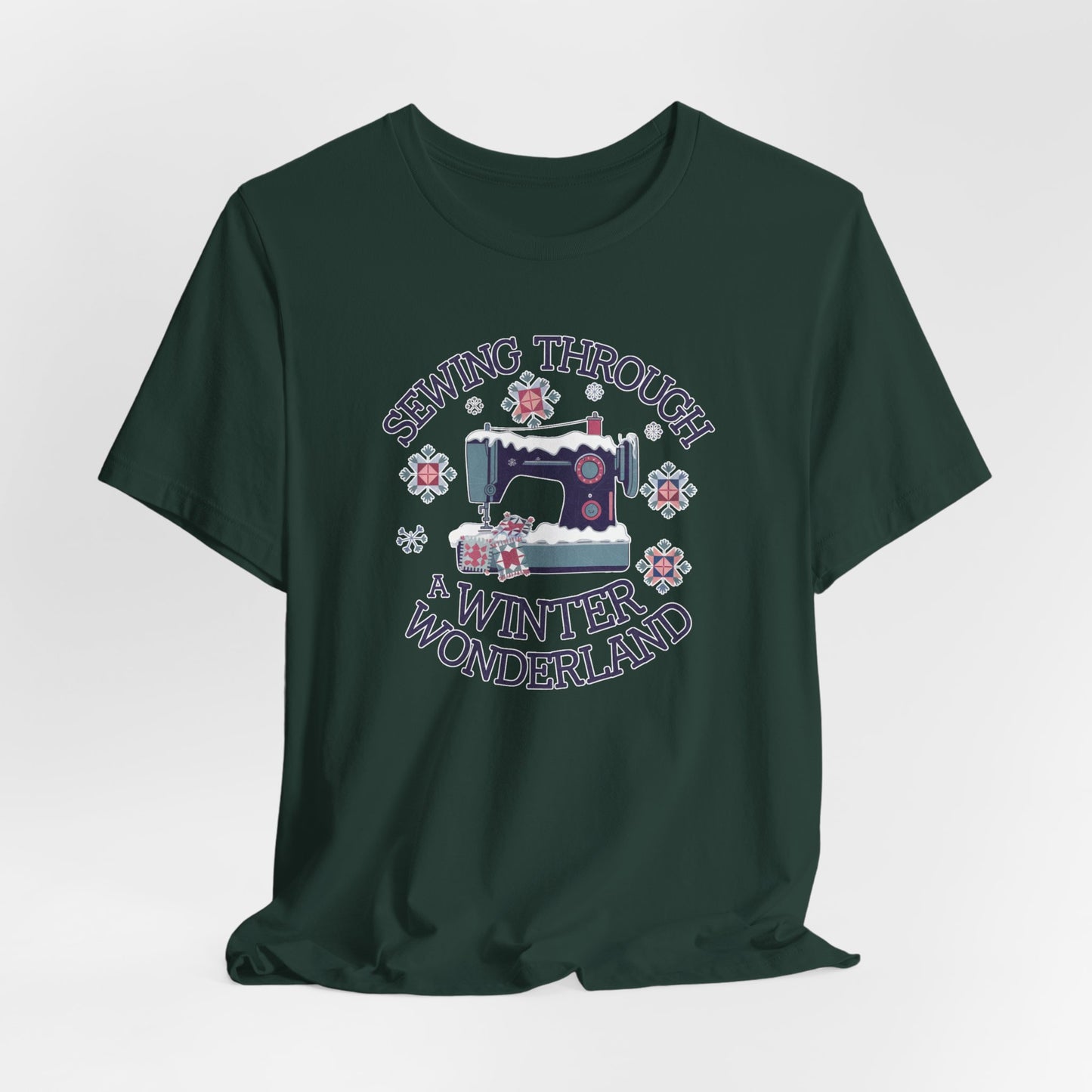 A Forest winter quilting t-shirt featuring a sewing machine covered in snow, surrounded by quilted snowflakes, with the phrase "Sewing Through a Winter Wonderland" above and below the image.