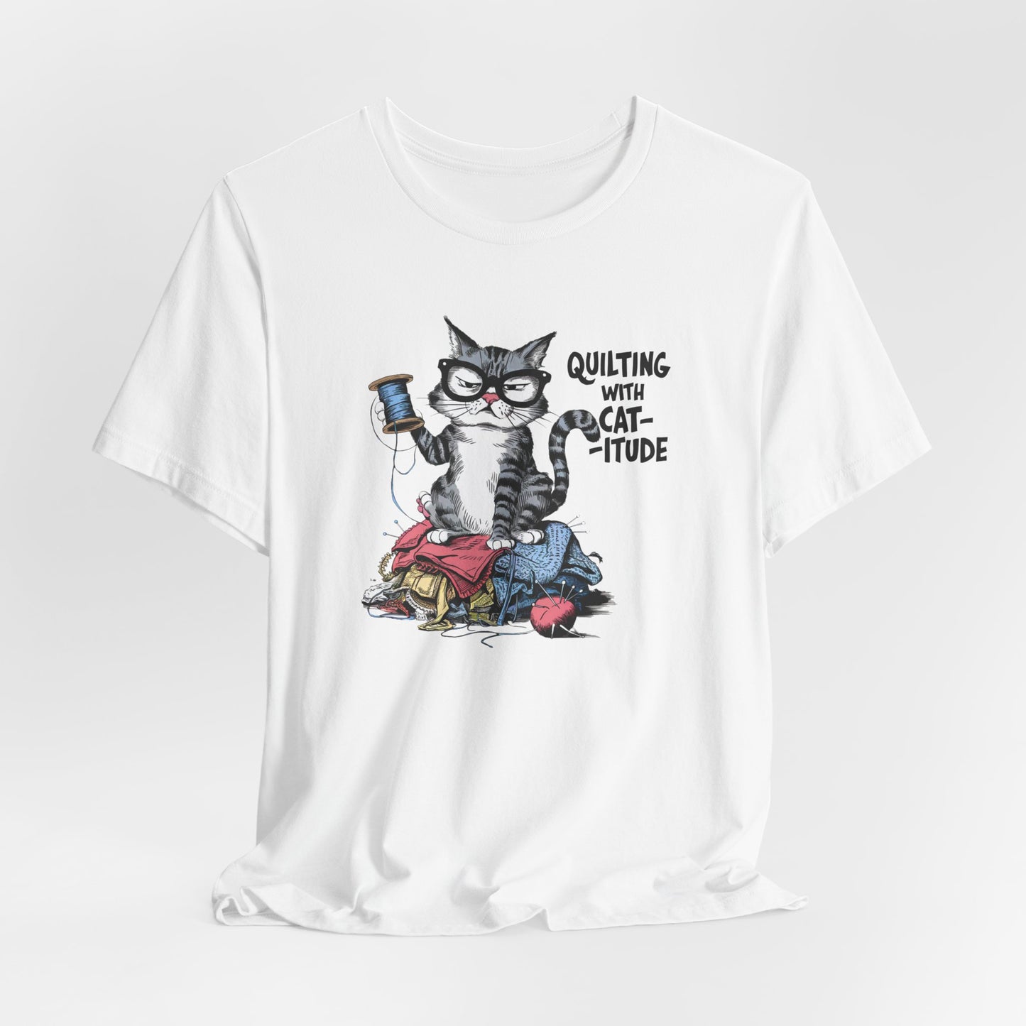 A White t-shirt featuring a sassy cartoon cat sitting on a pile of colorful fabric, holding a spool of thread, with the phrase 'Quilting with Cat-itude,' perfect for quilters and cat lovers.