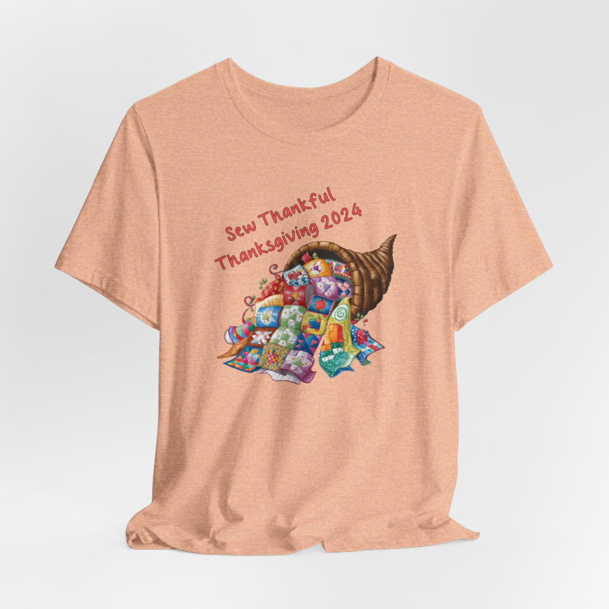 A Heather Peach thanksgiving quilting t-shirt featuring a colorful quilted cornucopia overflowing with various quilt patterns and designs. Text reads "Sew Thankful Thanksgiving 2024" in red. Vibrant illustration combines quilting motifs with Thanksgiving imagery.