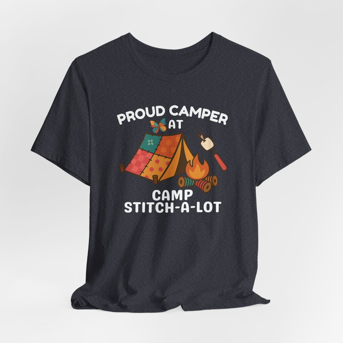 A A Heather Navy t-shirt featuring a colorful design with a patchwork tent, thread spool campfire, marshmallow on a seam ripper stick, and a butterfly. Text reads "Proud Camper at Camp Stitch-a-Lot." Fun gift for quilting retreats.