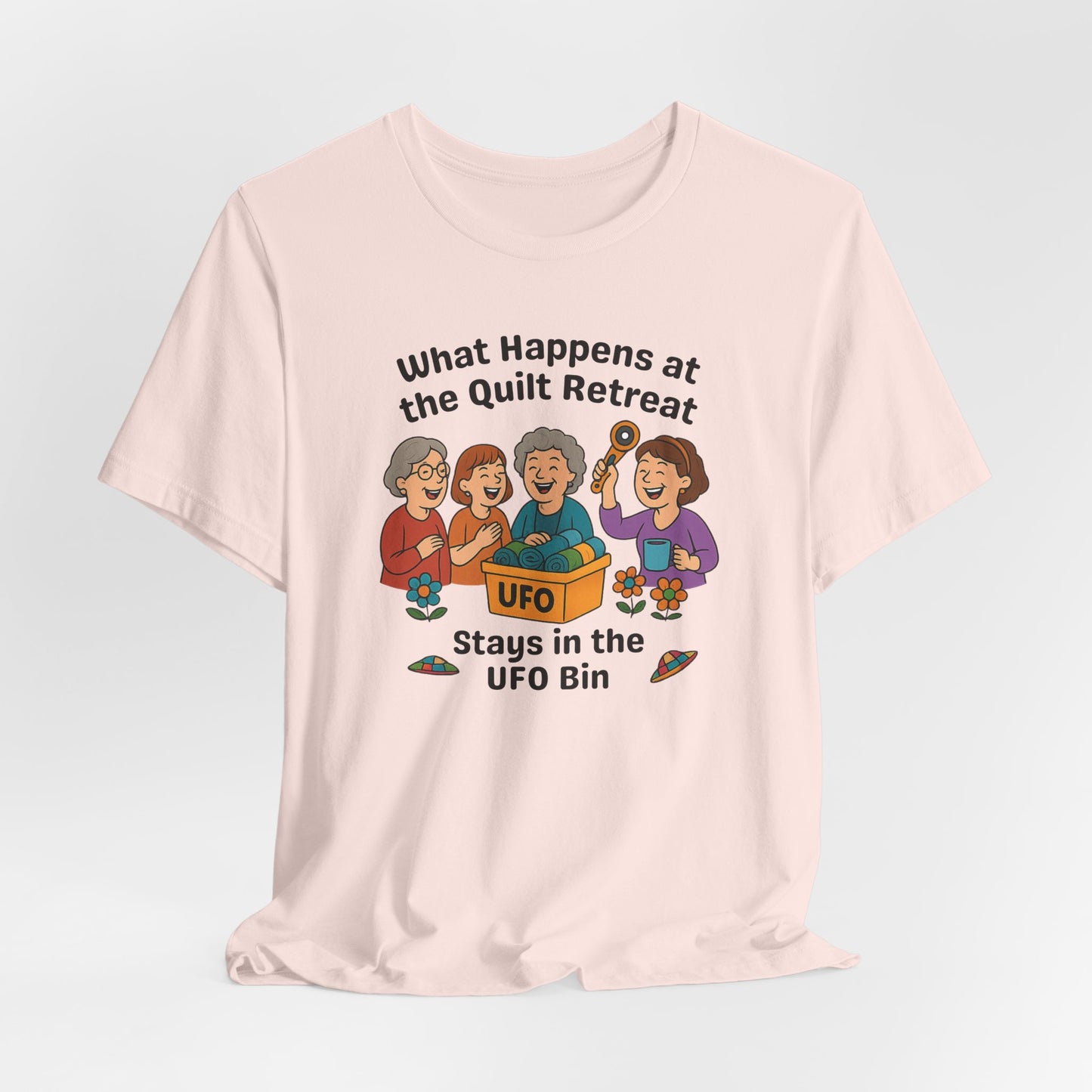 A A Soft Pink t-shirt featuring a cartoon design of four laughing women at a quilt retreat with a bin labeled “UFO” full of fabric. Surrounded by patchwork flying saucers and flowers. Text reads “What happens at the quilt retreat stays in the UFO bin.”