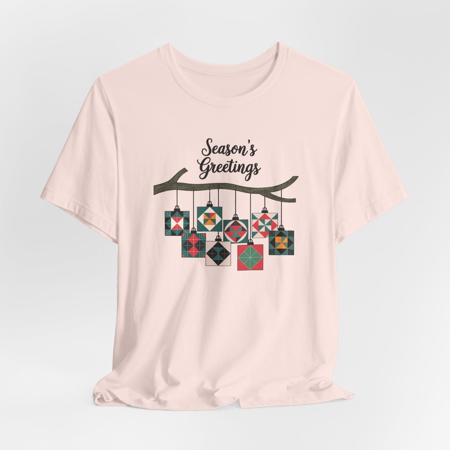 A Soft Pink christmas quilting t-shirt featuring quilt block ornaments hanging from a branch, featuring the phrase "Season's Greetings" above. The ornaments showcase various quilting patterns to highlight the holiday quilting theme.